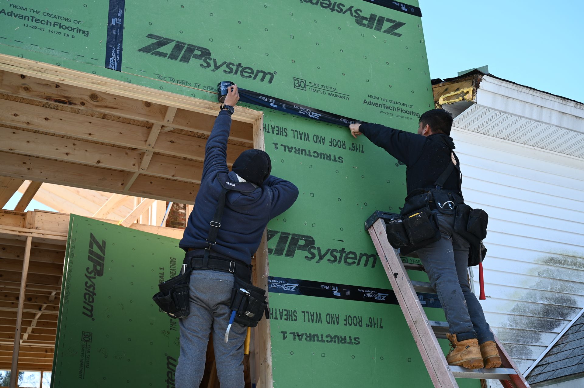 Two workers in dark clothing and tool belts applying black tape to the seams of green ZIP System wall panels on a house.