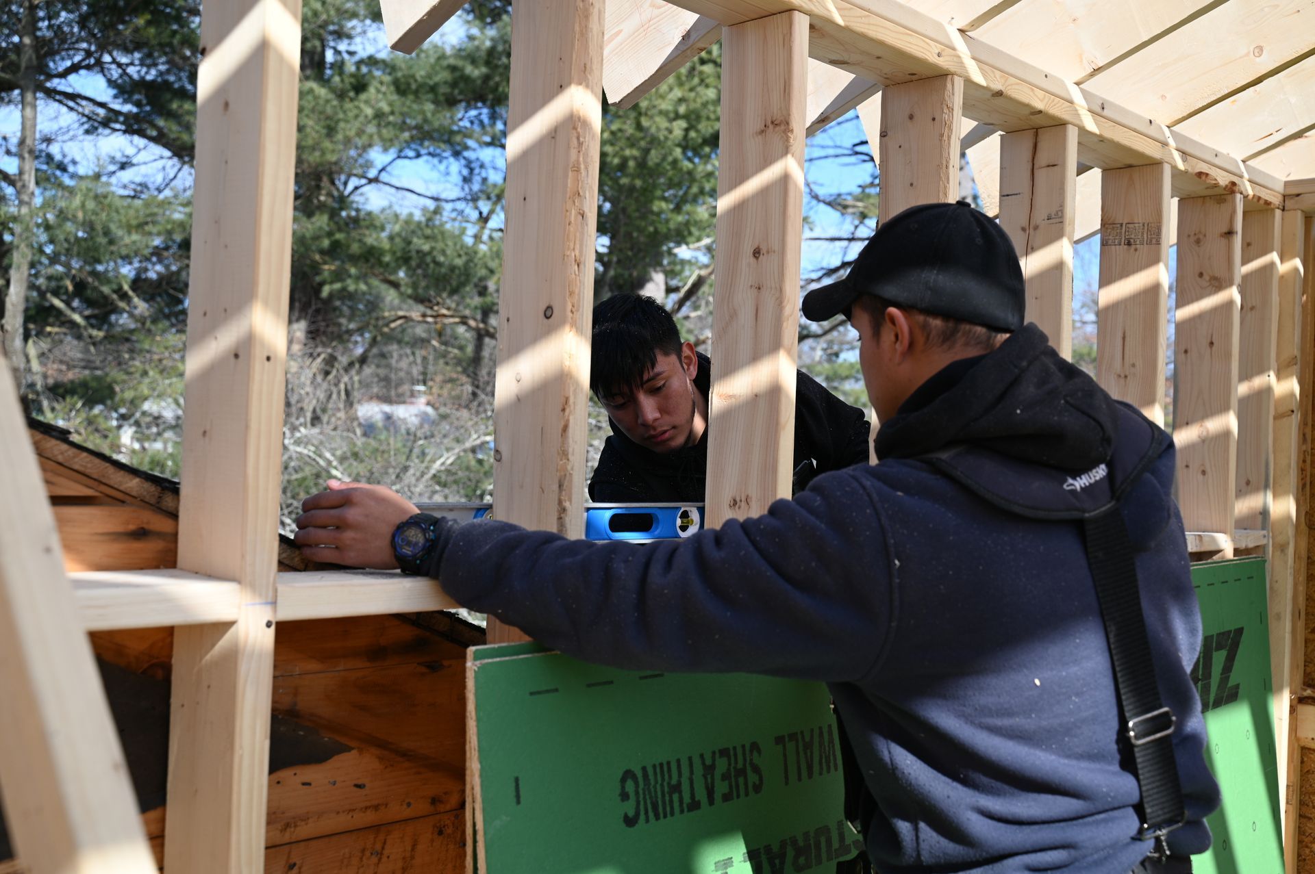 Two people work on framing a wooden building structure with green wall sheathing outdoors.