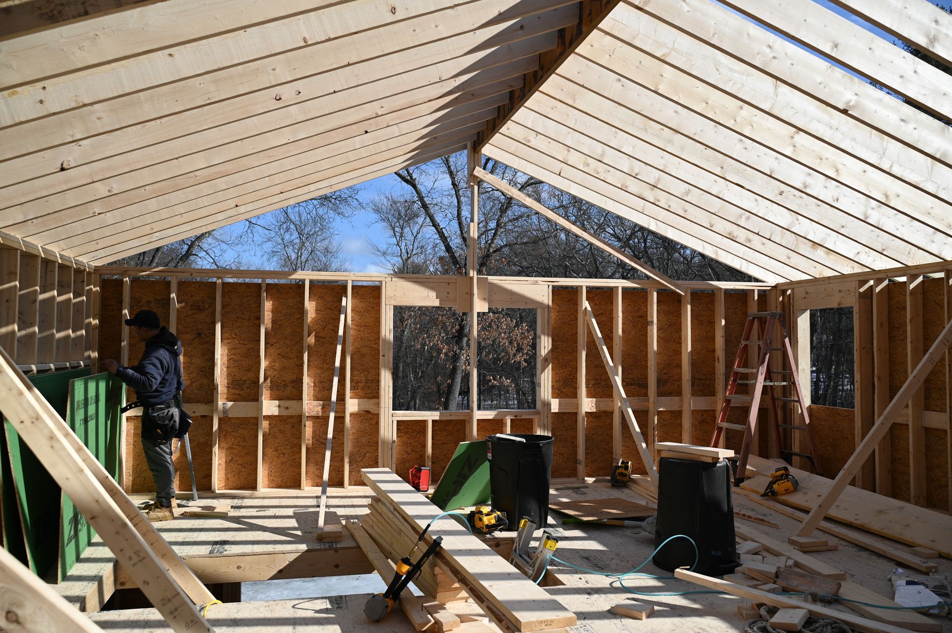 A person in a dark jacket works on the wooden framing of a house under construction with exposed rafters.