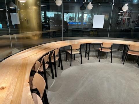 A curved wooden counter with light-colored chairs in a modern, glass-walled cafe space.