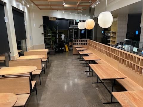 Modern cafe interior with light wood tables, bench seating, pendant globe lighting, and a polished concrete floor.