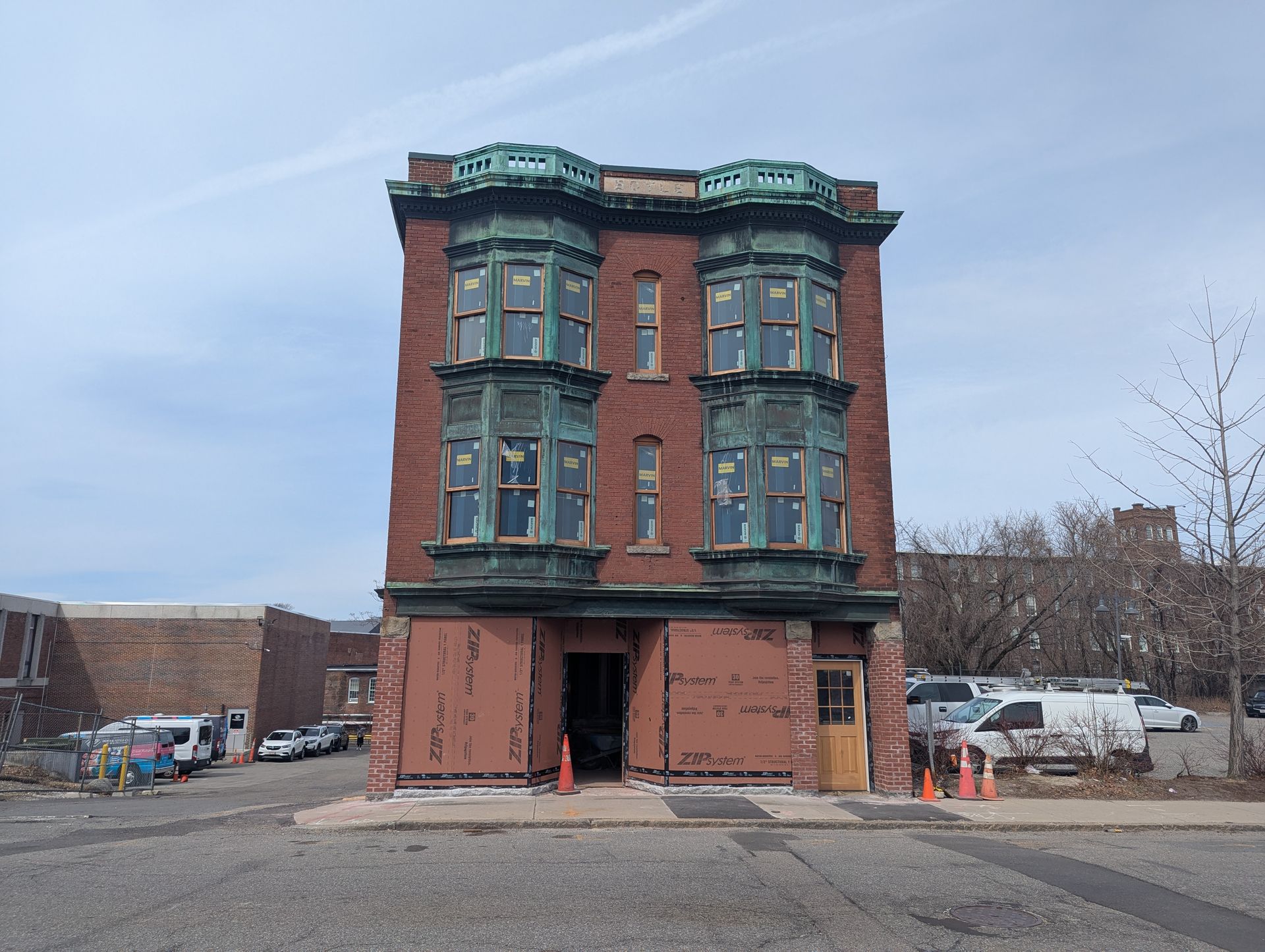 A three-story brick building with copper-clad bay windows under construction, situated in a paved urban lot.