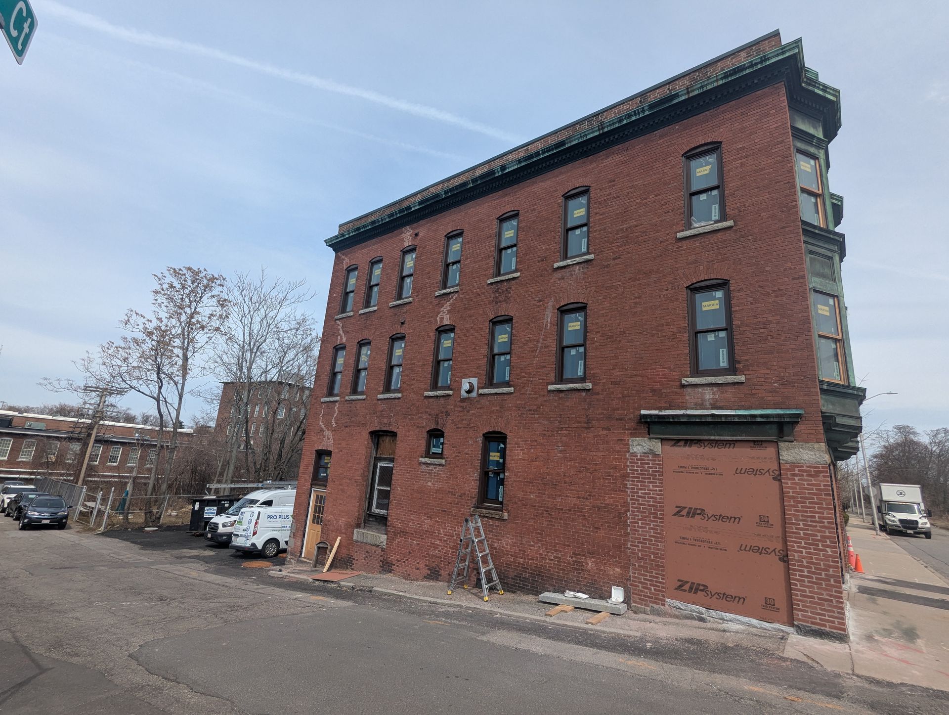 A four-story red brick building on a street corner with new black-framed windows, construction equipment, and parked cars.