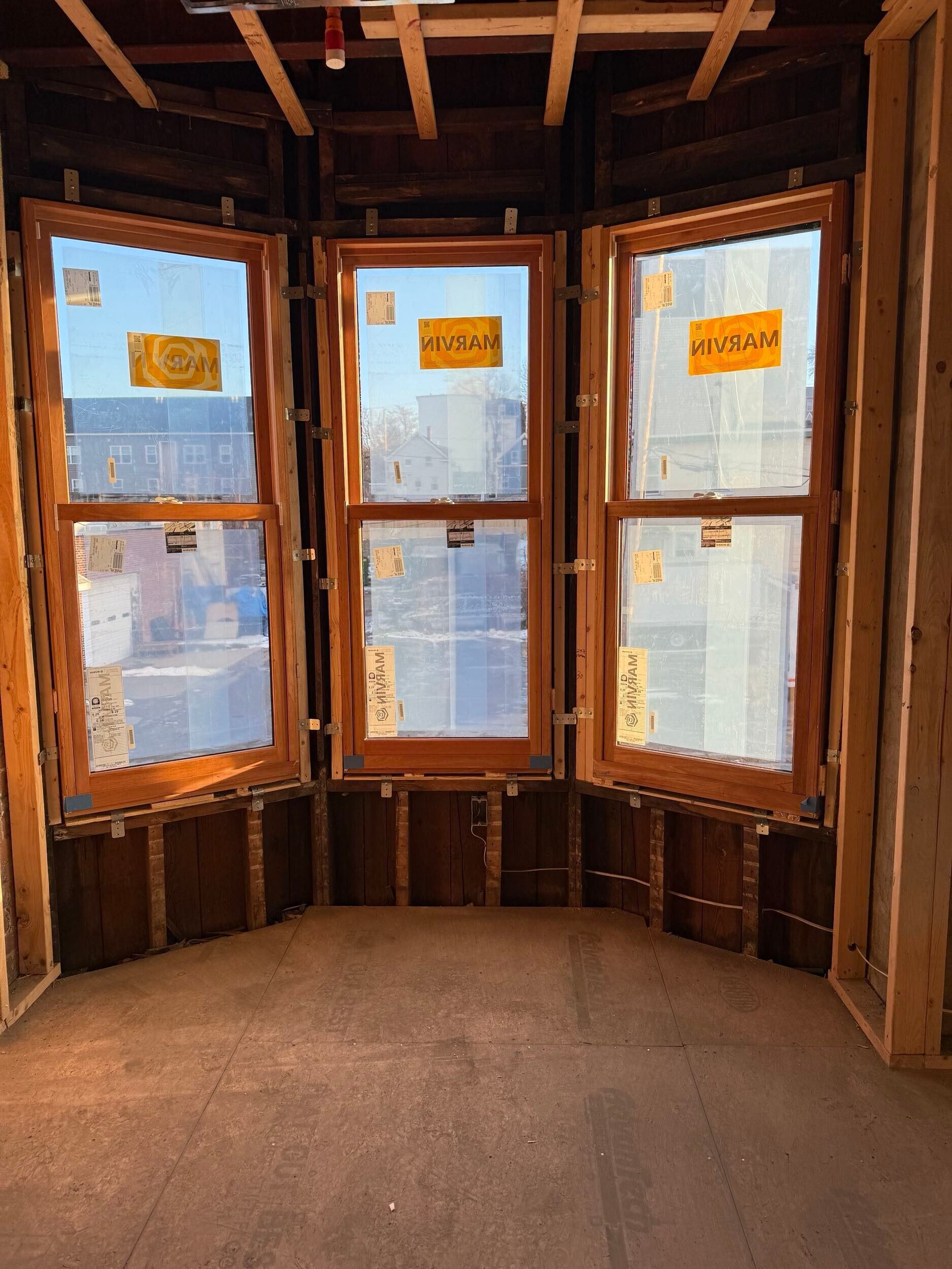 Three wooden-framed windows installed in a bay wall under construction with exposed wooden studs and subflooring.
