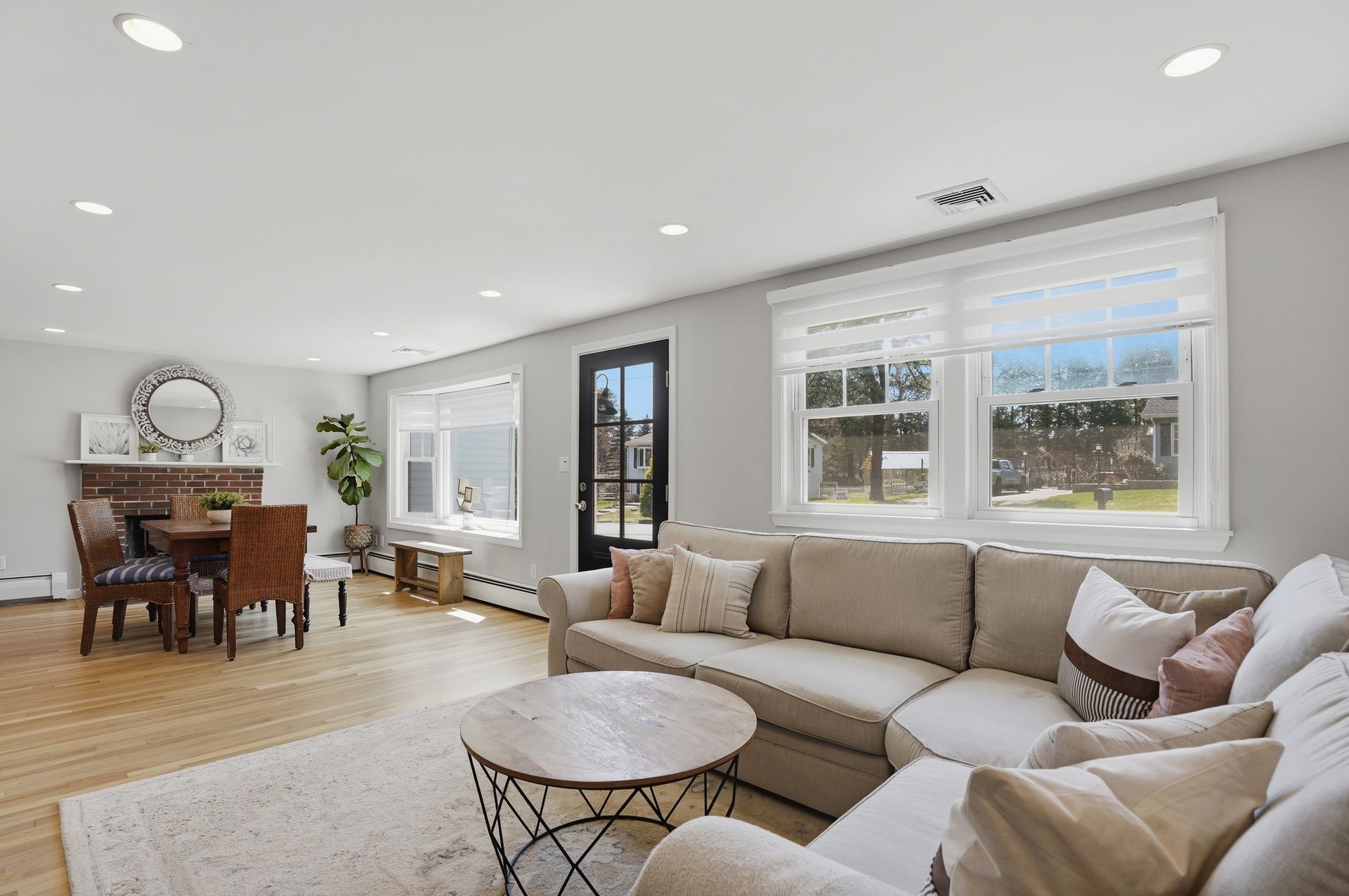 Bright open living room with beige sectional, wooden dining table, large windows, and light hardwood floors