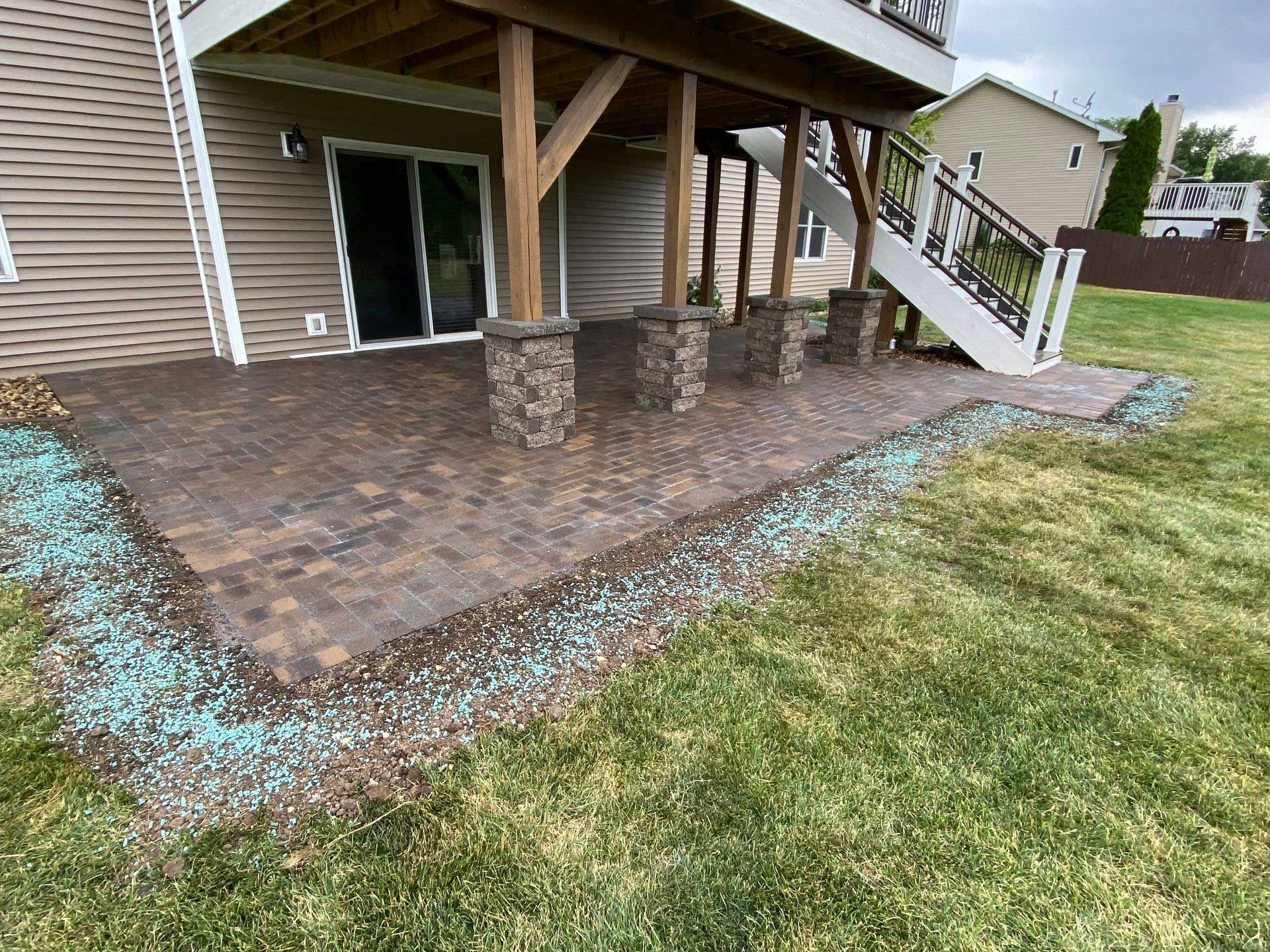 A patio under a deck in a backyard with a house in the background.
