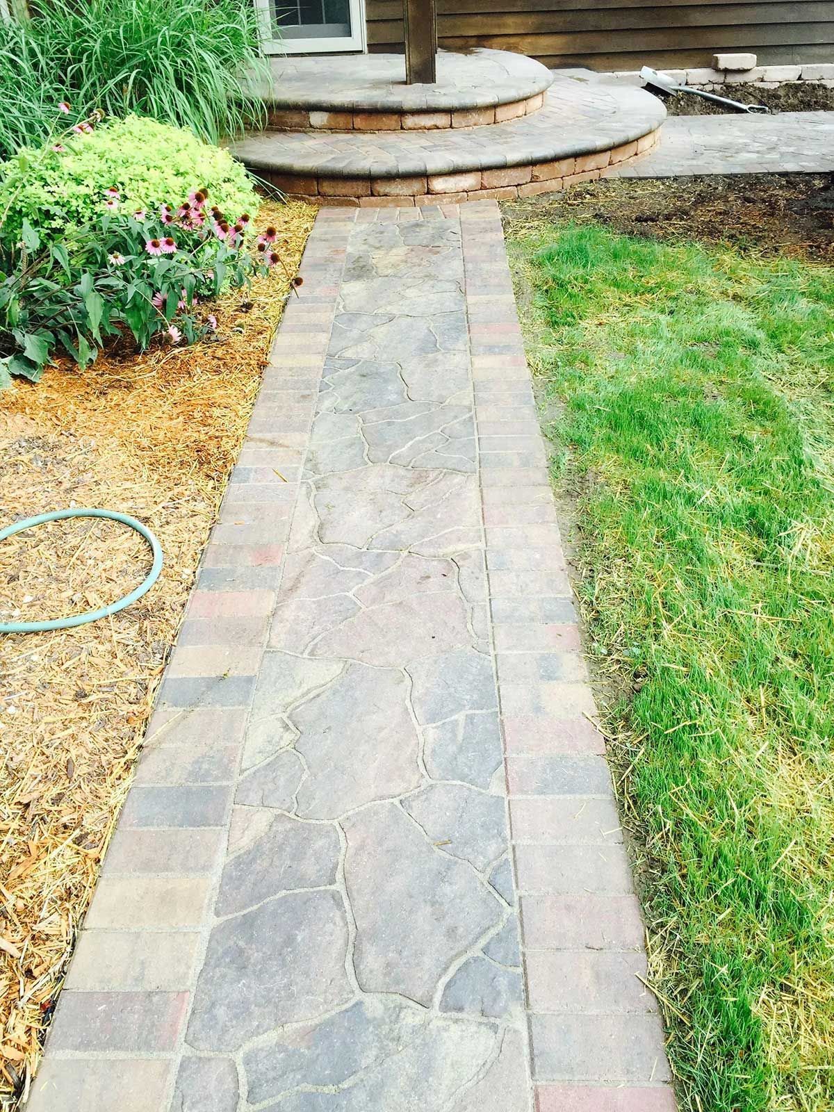 A brick walkway leading to a house with a hose on the side of it.