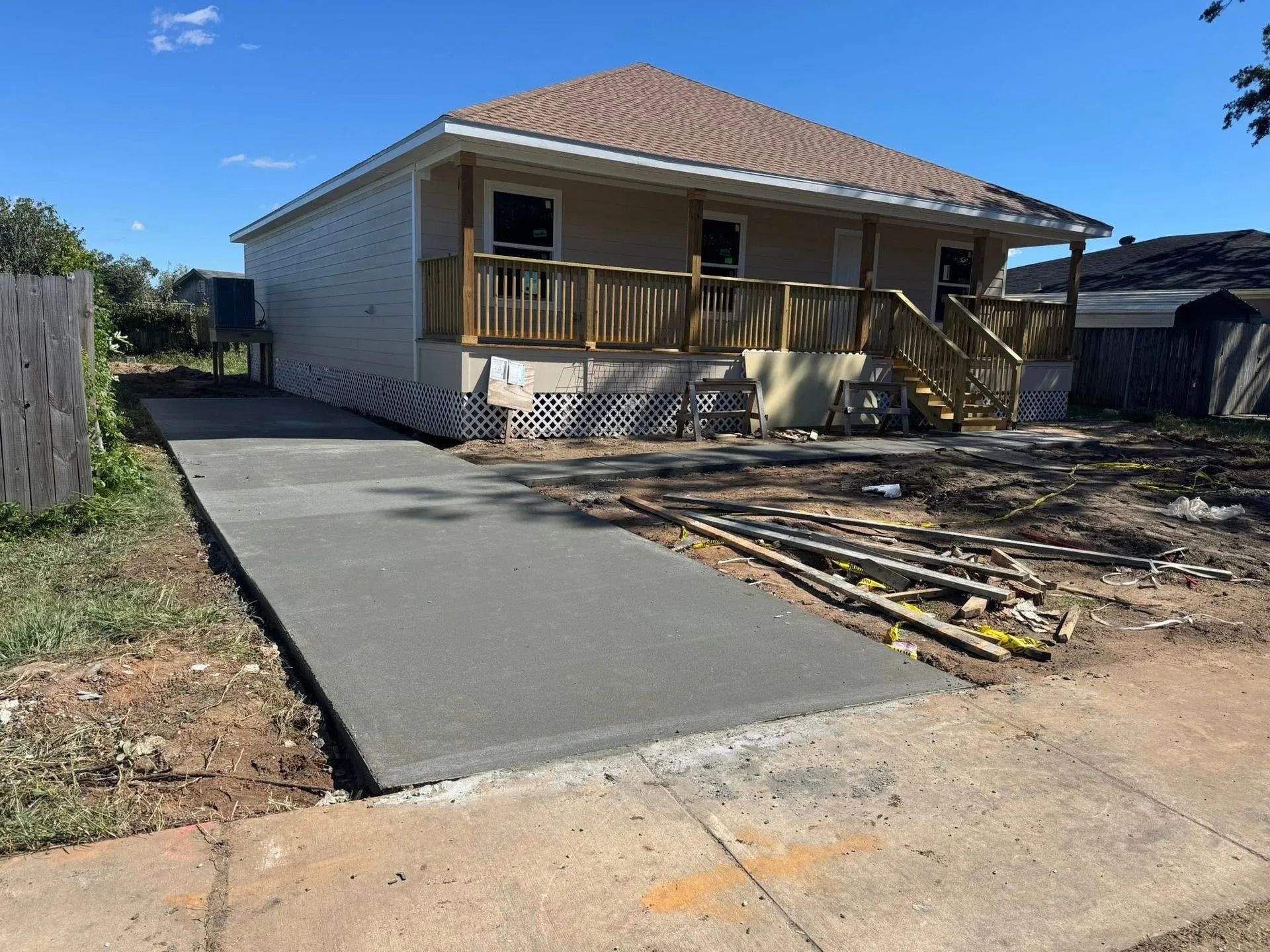 Newly poured concrete driveway leading to a beige house with a wooden porch and brown roof under a blue sky.