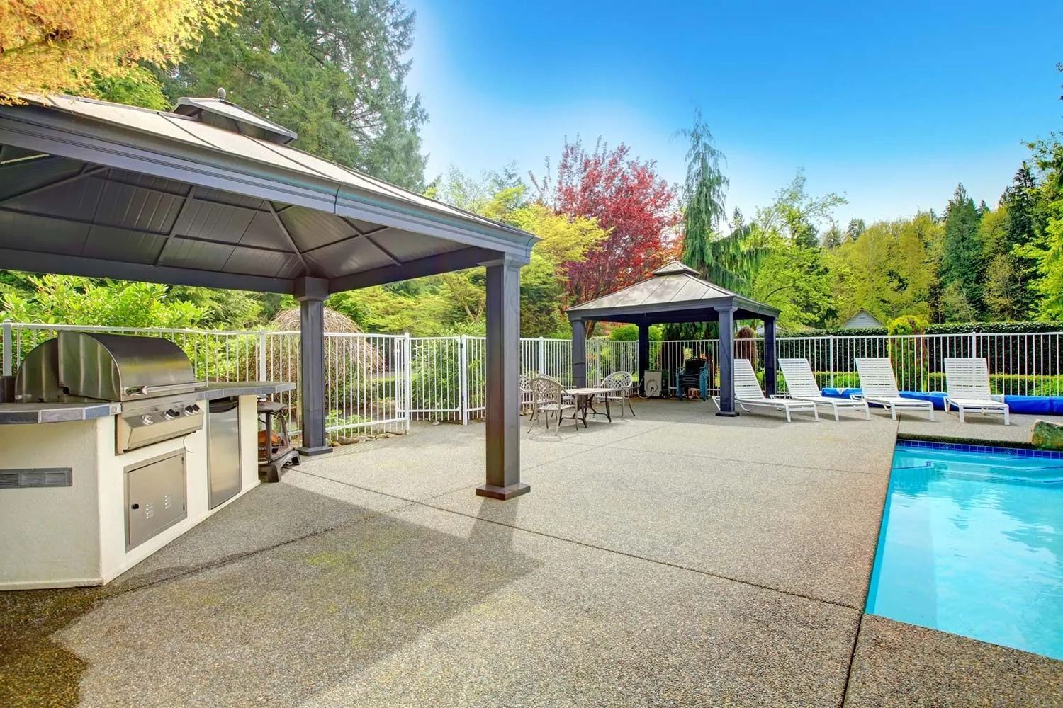 Outdoor patio with a pool, grill, and two gazebos under a blue sky and trees.