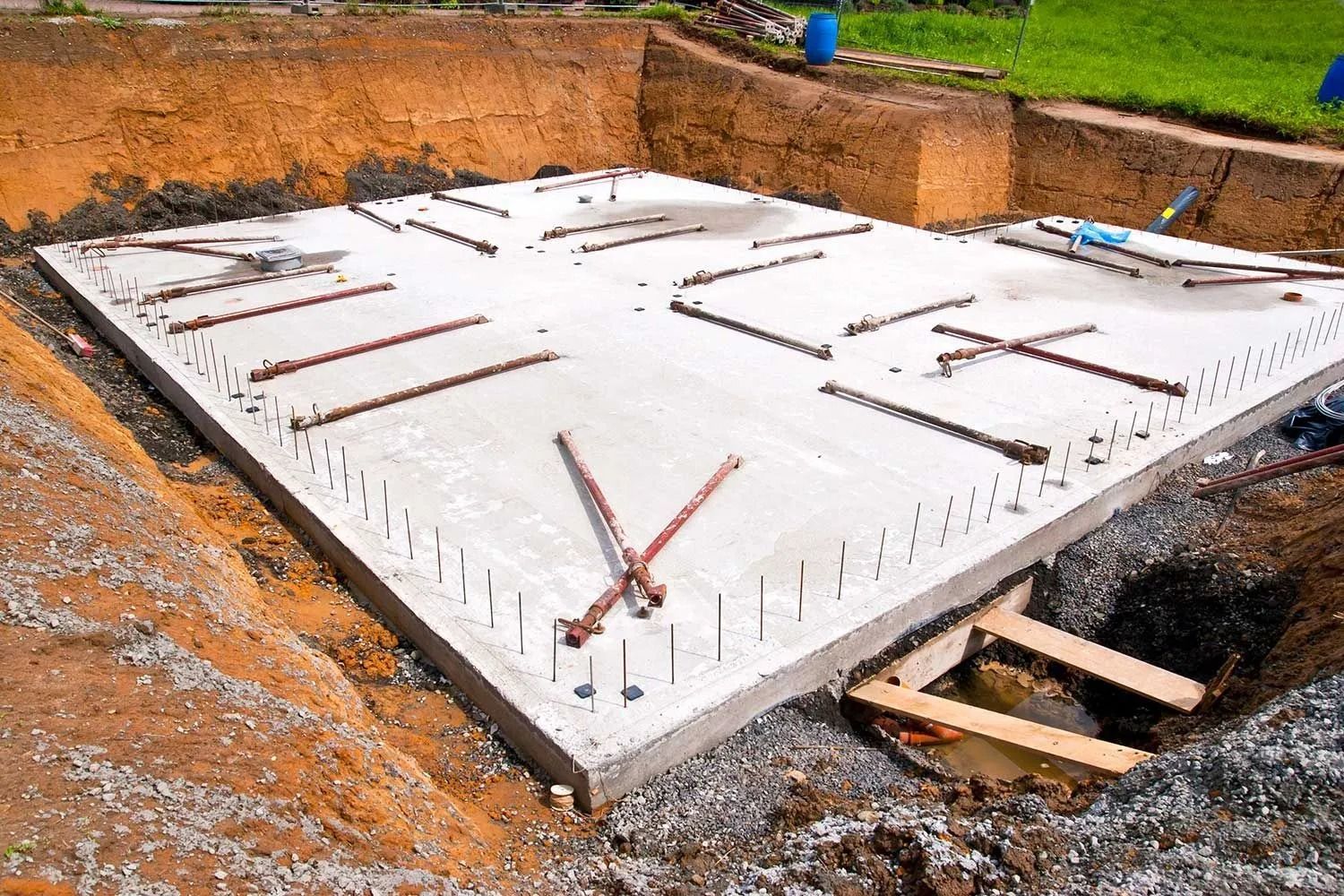 Concrete slab foundation under construction in an excavated area with rebar framework and wooden supports.