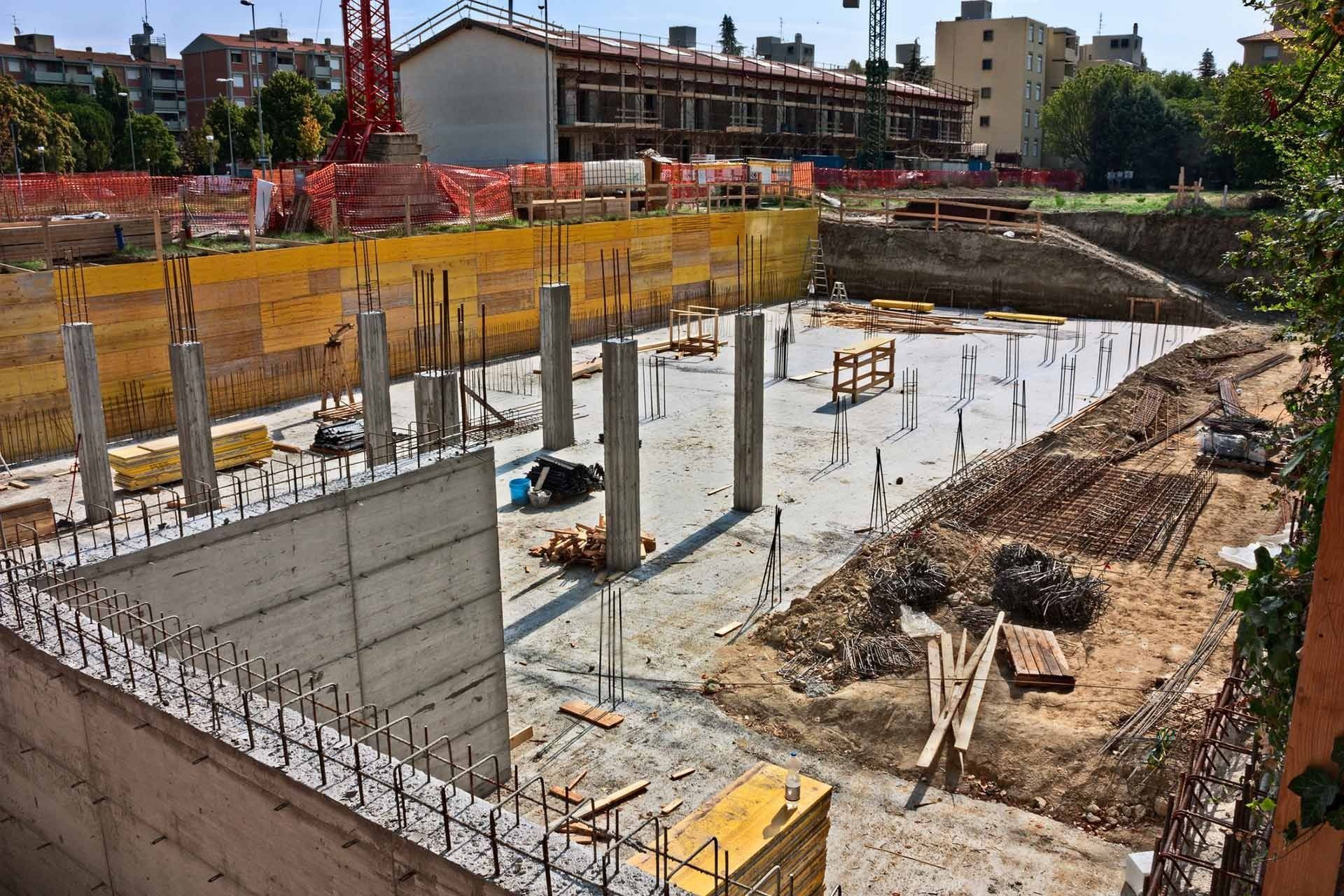 Construction site with exposed concrete foundation, pillars, and retaining walls, surrounded by fencing.