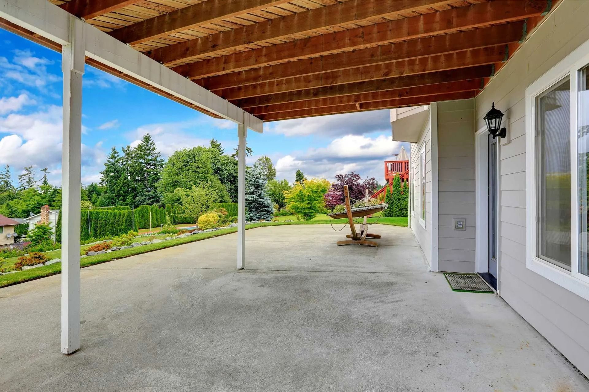 Covered patio with a concrete floor, overlooking a yard with trees and a blue sky.