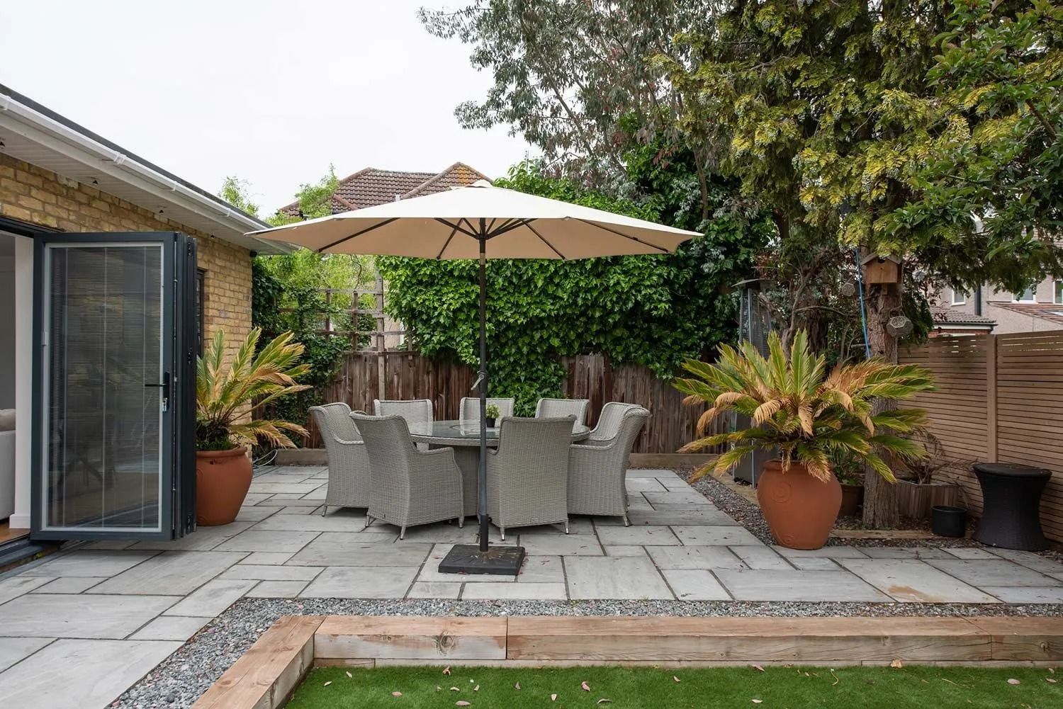 Patio with dining set, umbrella, and potted plants next to a house with open doors, under a cloudy sky.