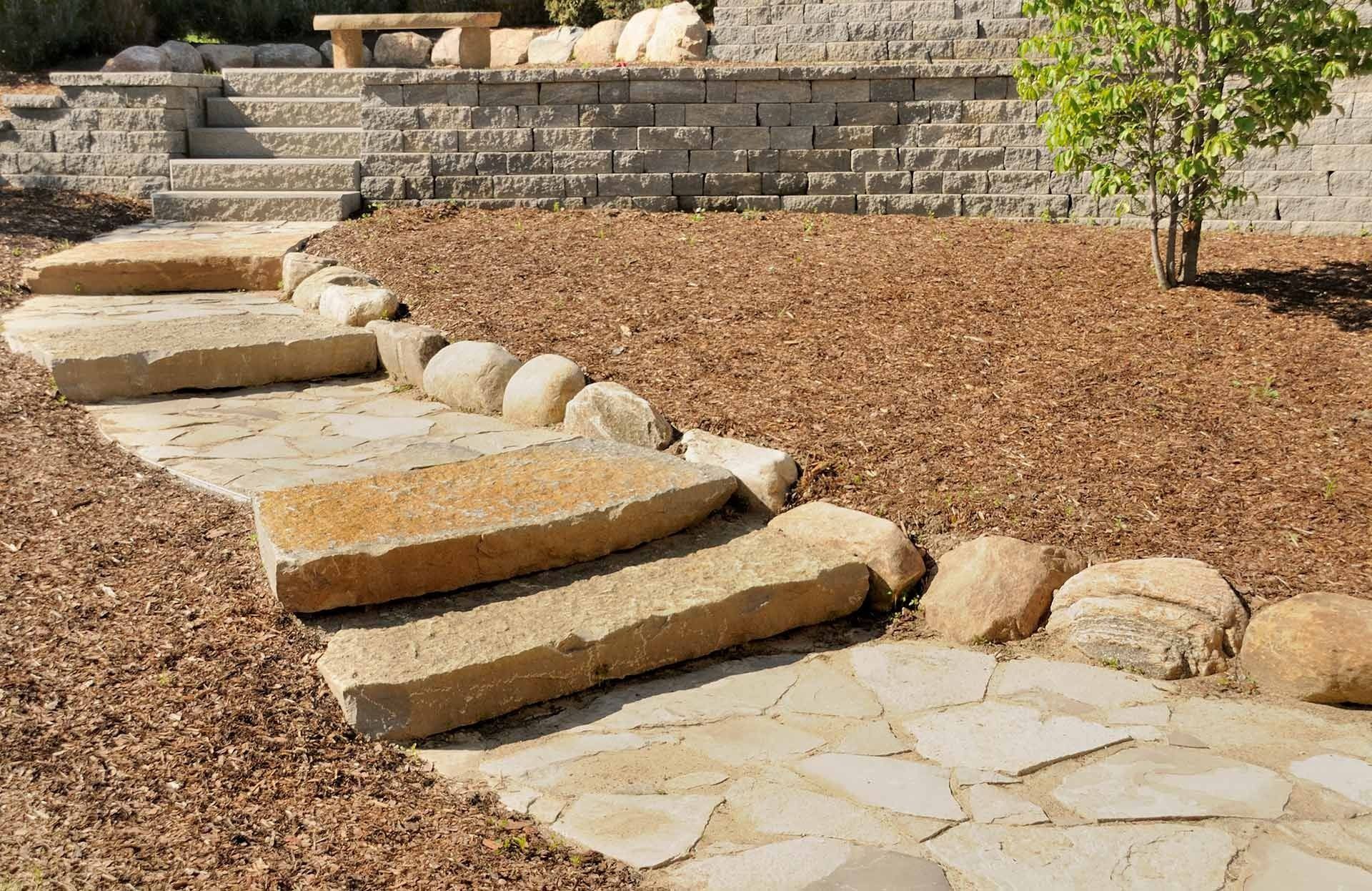 Stone pathway and steps lead up a landscaped yard, mulch and tree present.
