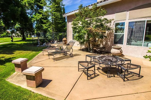 Patio with outdoor furniture, including a fire pit, next to a beige house, green lawn and trees.