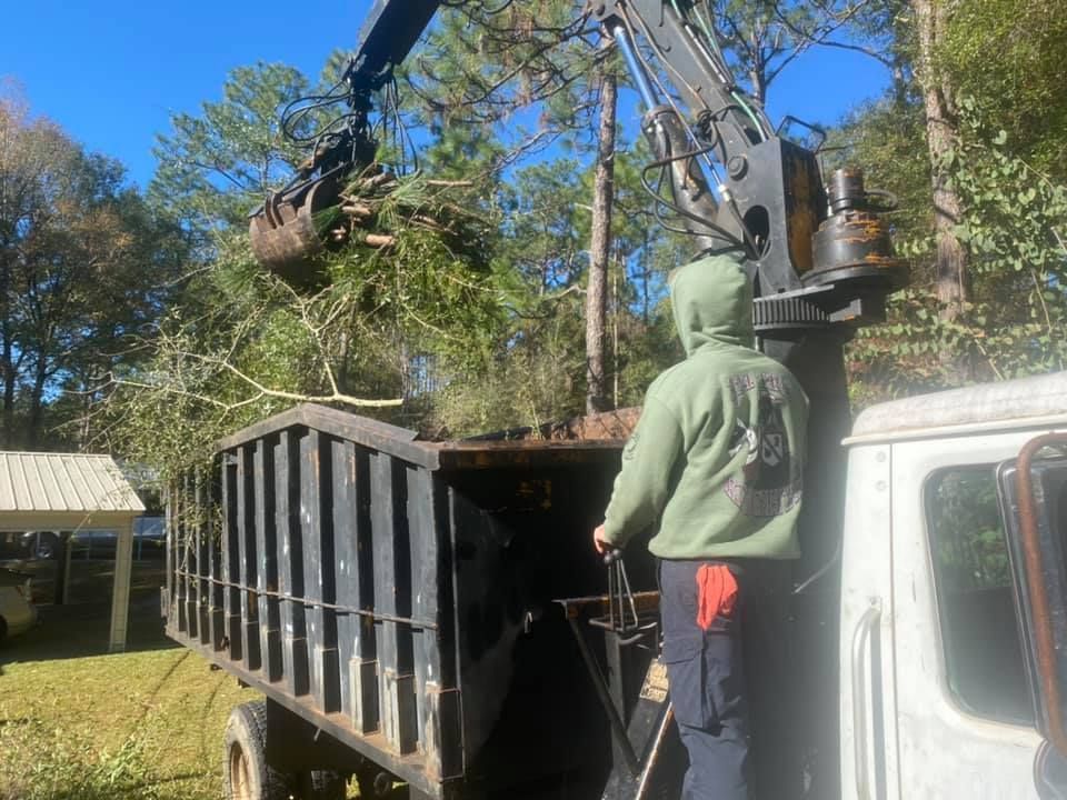 Man loading tree branches into a truck bed with a crane, outdoors on a sunny day.
