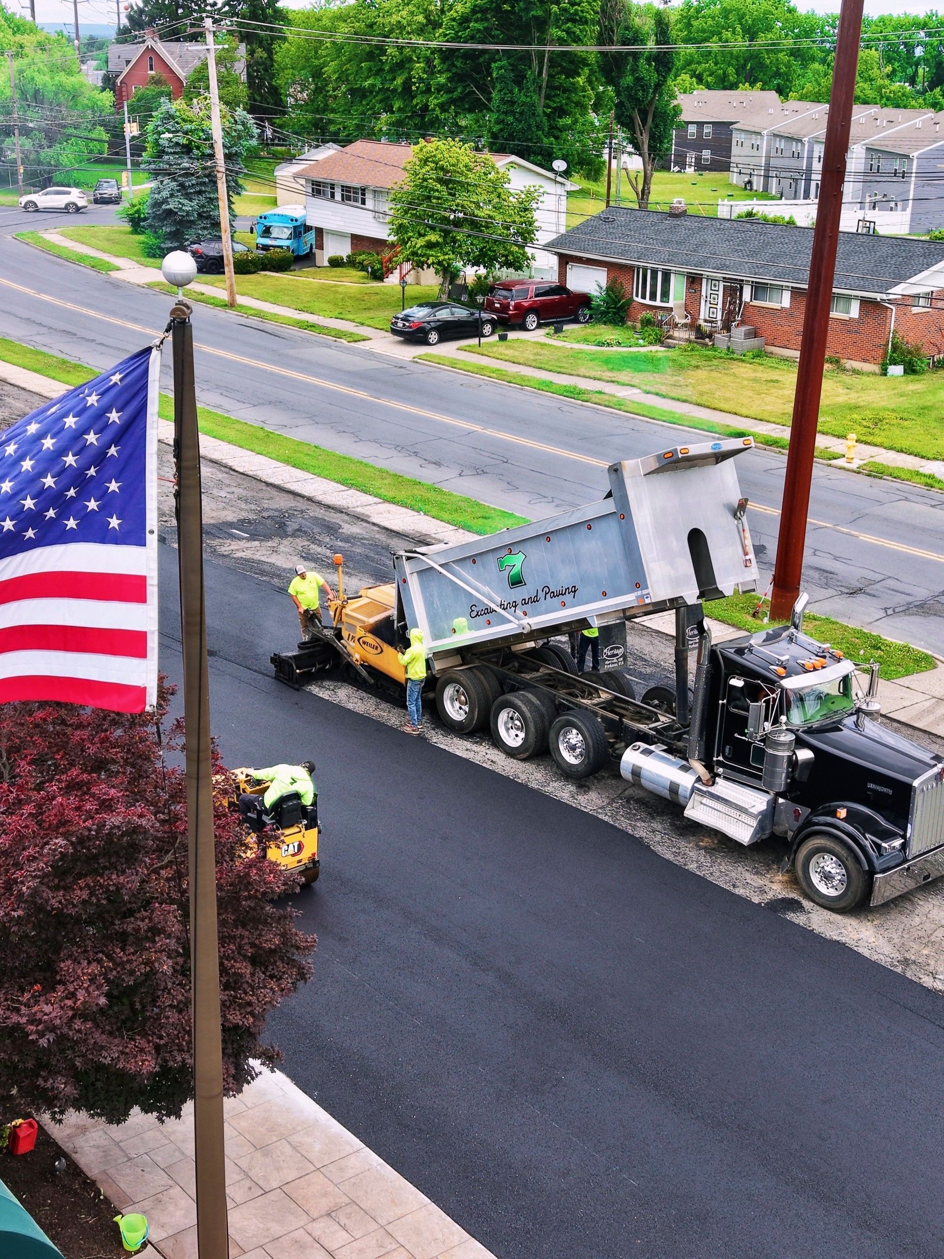 Flatbed truck hauling a gray boat on a suburban street beside an American flag and trees