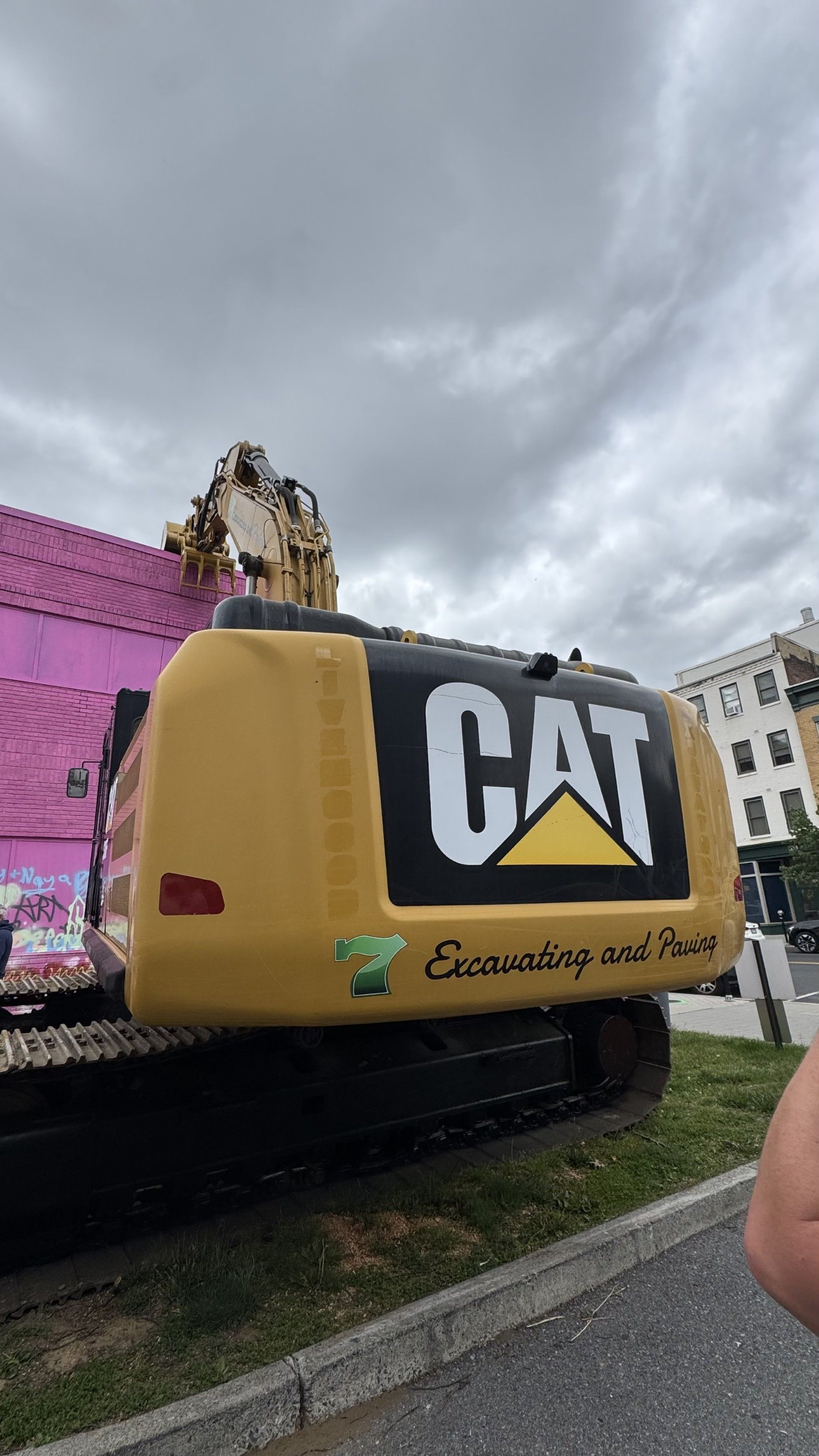 Yellow CAT excavator on a road, with a person standing on top and pink construction barriers behind