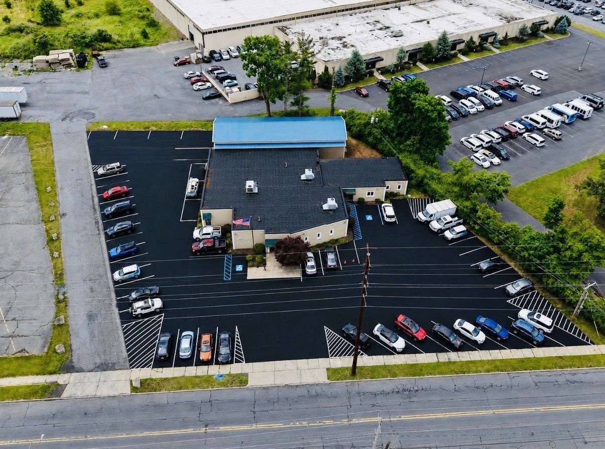 Aerial view of a small building with a blue roof and parking lot filled with cars beside a road.