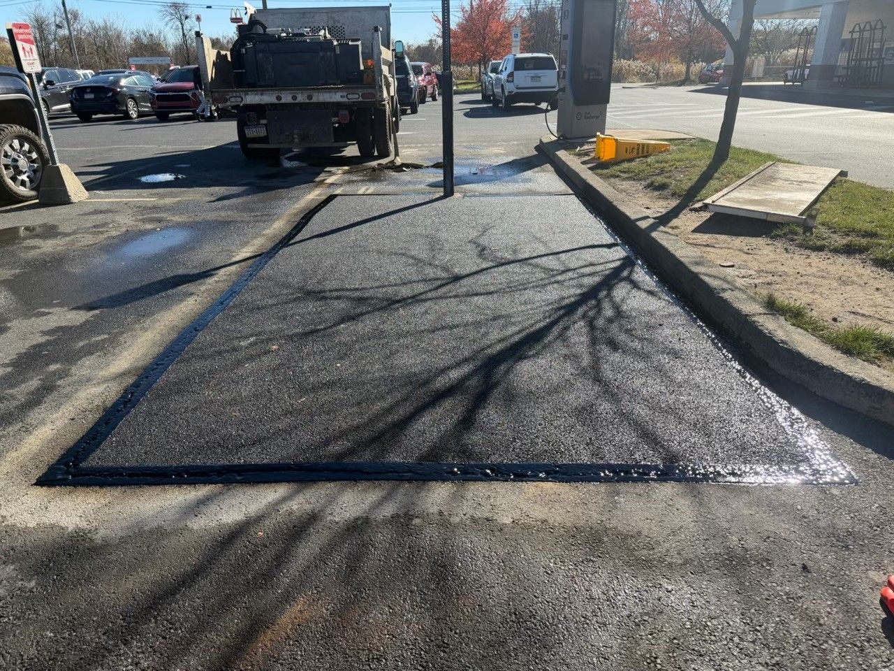 Freshly applied black asphalt patch in a parking lot, bordered by a curb and surrounding older pavement.