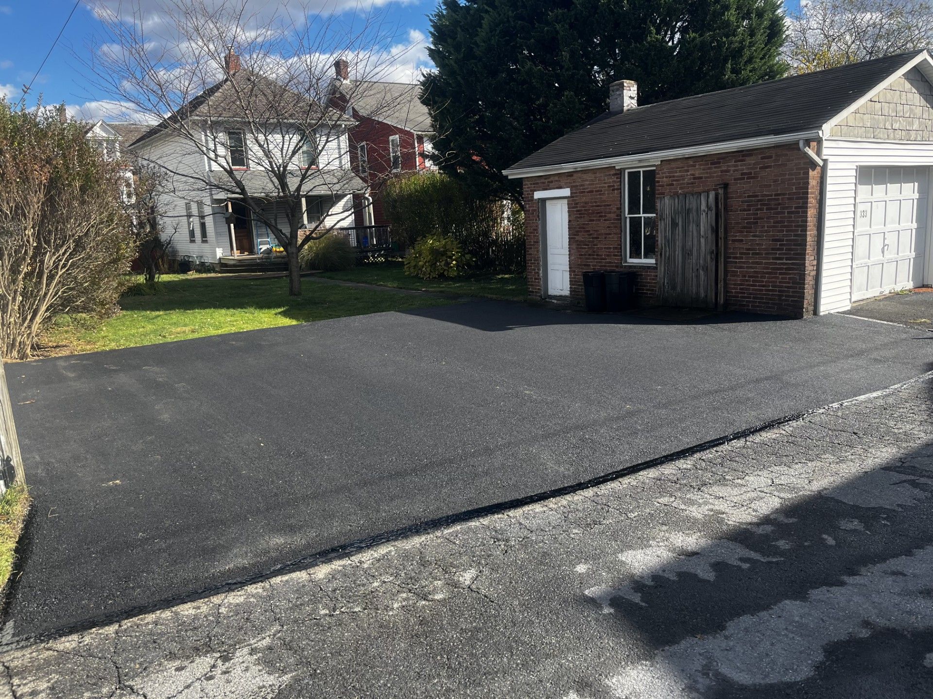 A newly paved, dark asphalt driveway leads to a brick garage next to a white house under a blue sky.