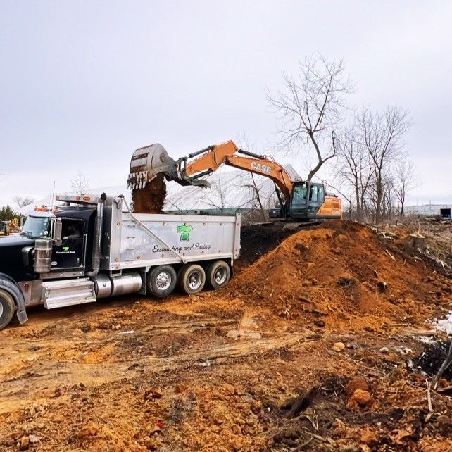 An orange excavator loading soil into the back of a dump truck at a construction site under an overcast sky.