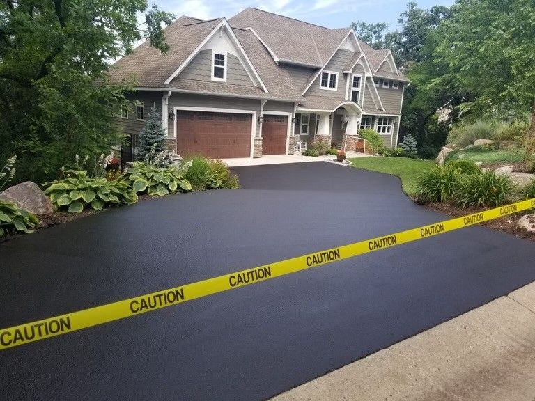 A freshly paved, black driveway in front of a two-story house, blocked by yellow caution tape.