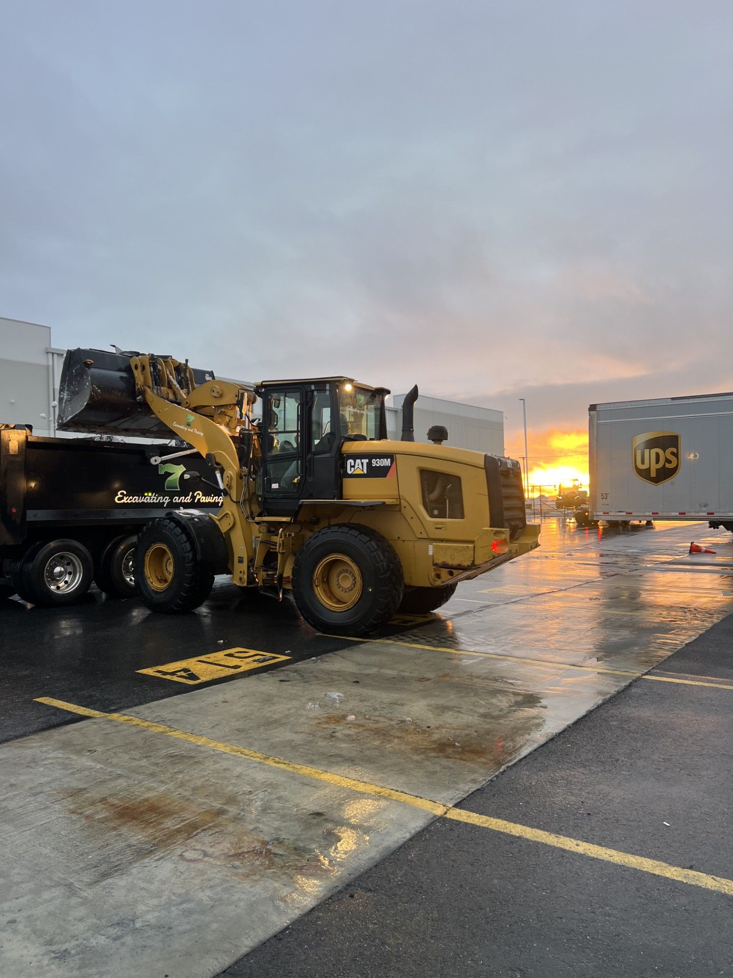 A yellow Caterpillar front loader fills the bed of a black dump truck at sunset near a parked UPS trailer.