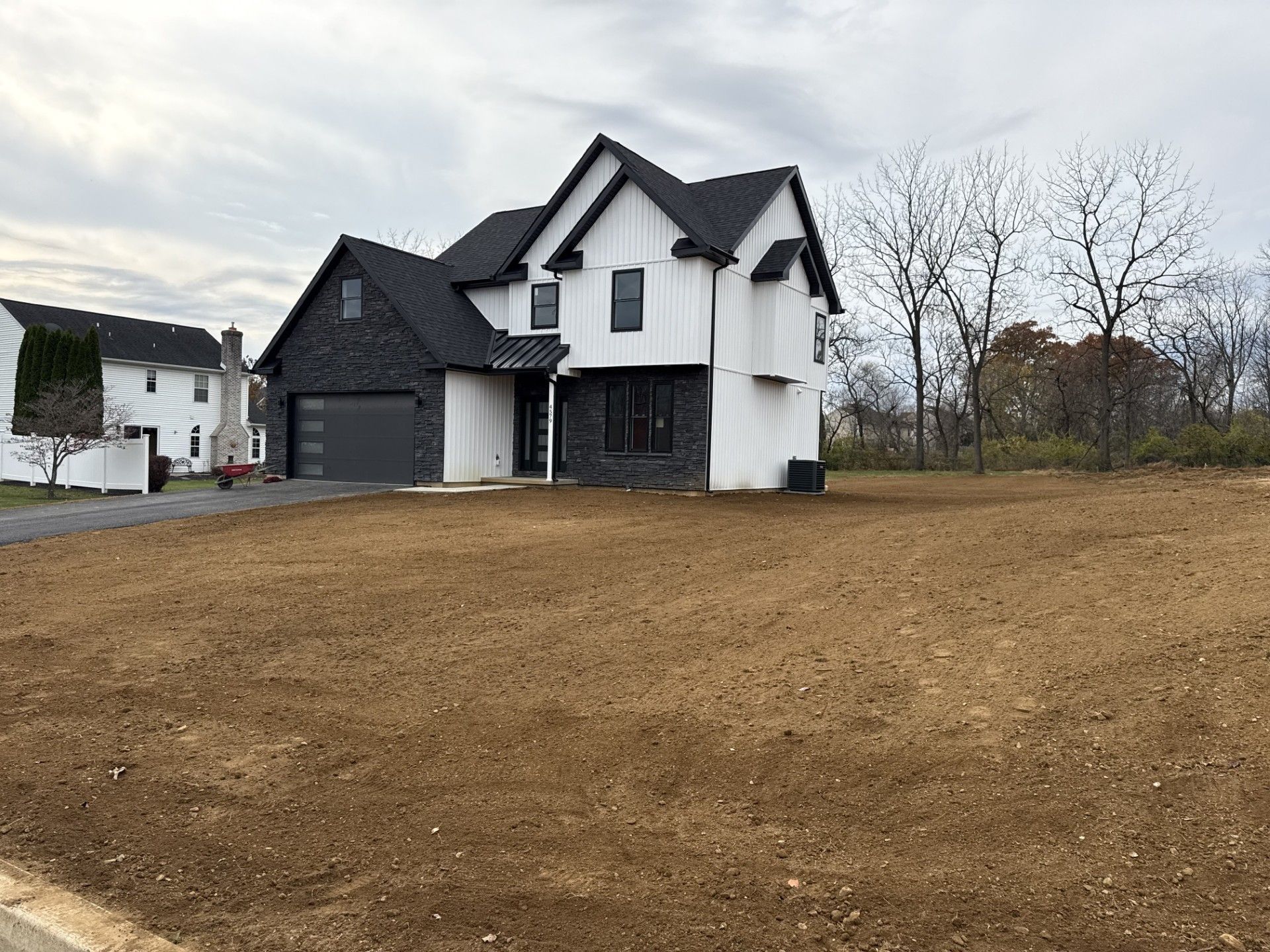 A newly constructed two-story house with dark stone and white siding stands on a large, empty, dirt-covered lawn.