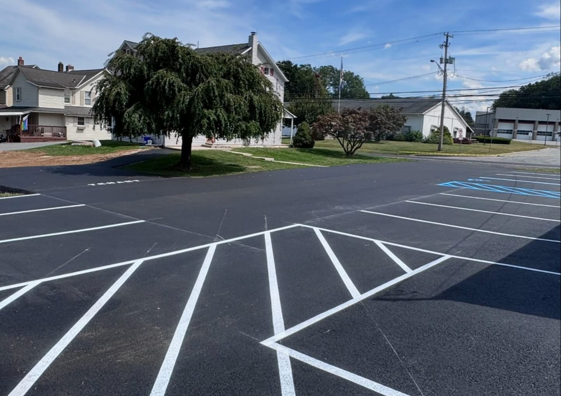 Newly paved parking lot with white lines, trees, and buildings under a blue sky.