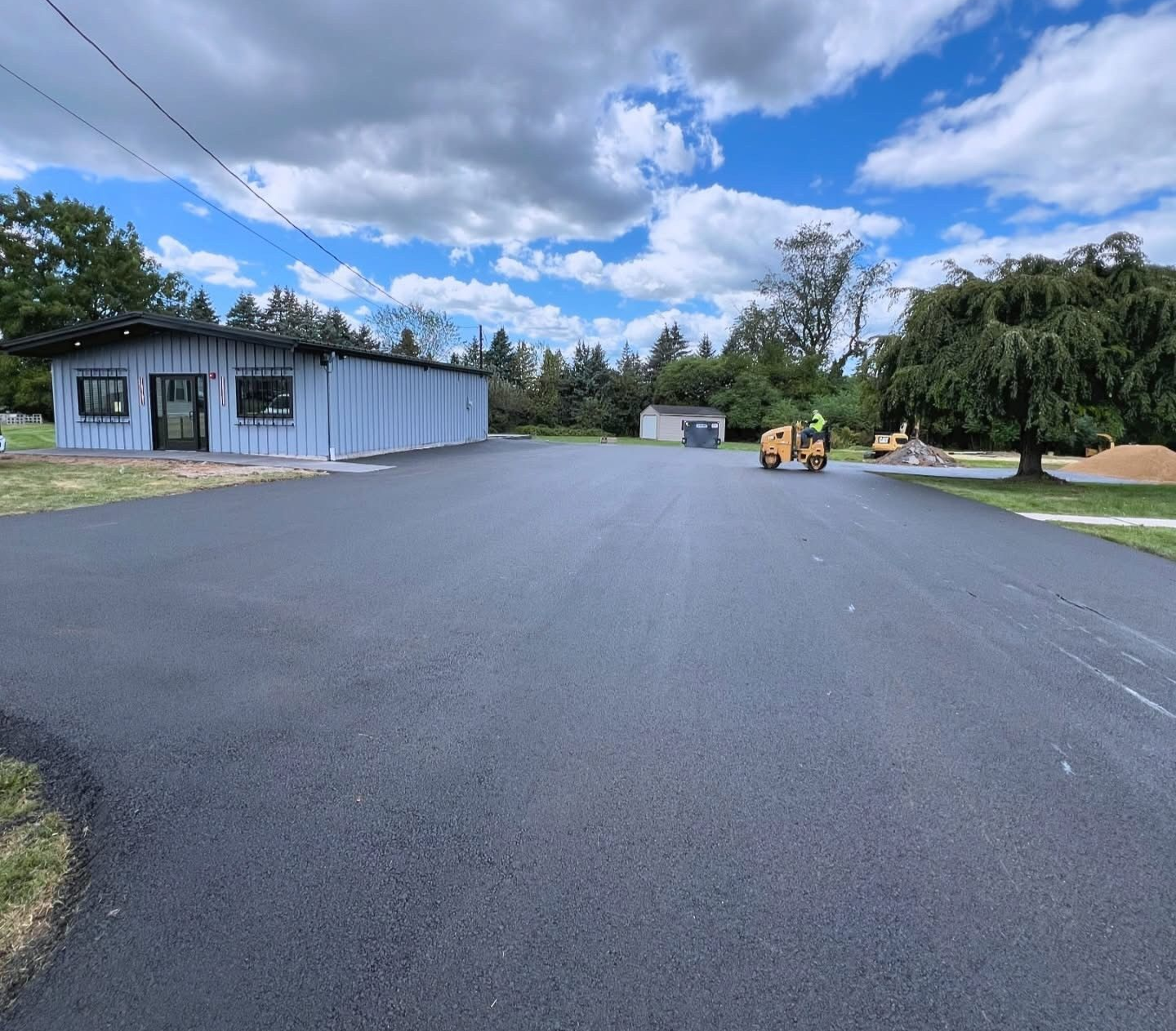 Black asphalt paved lot next to a light blue building under a cloudy sky. A small yellow machine is operating.