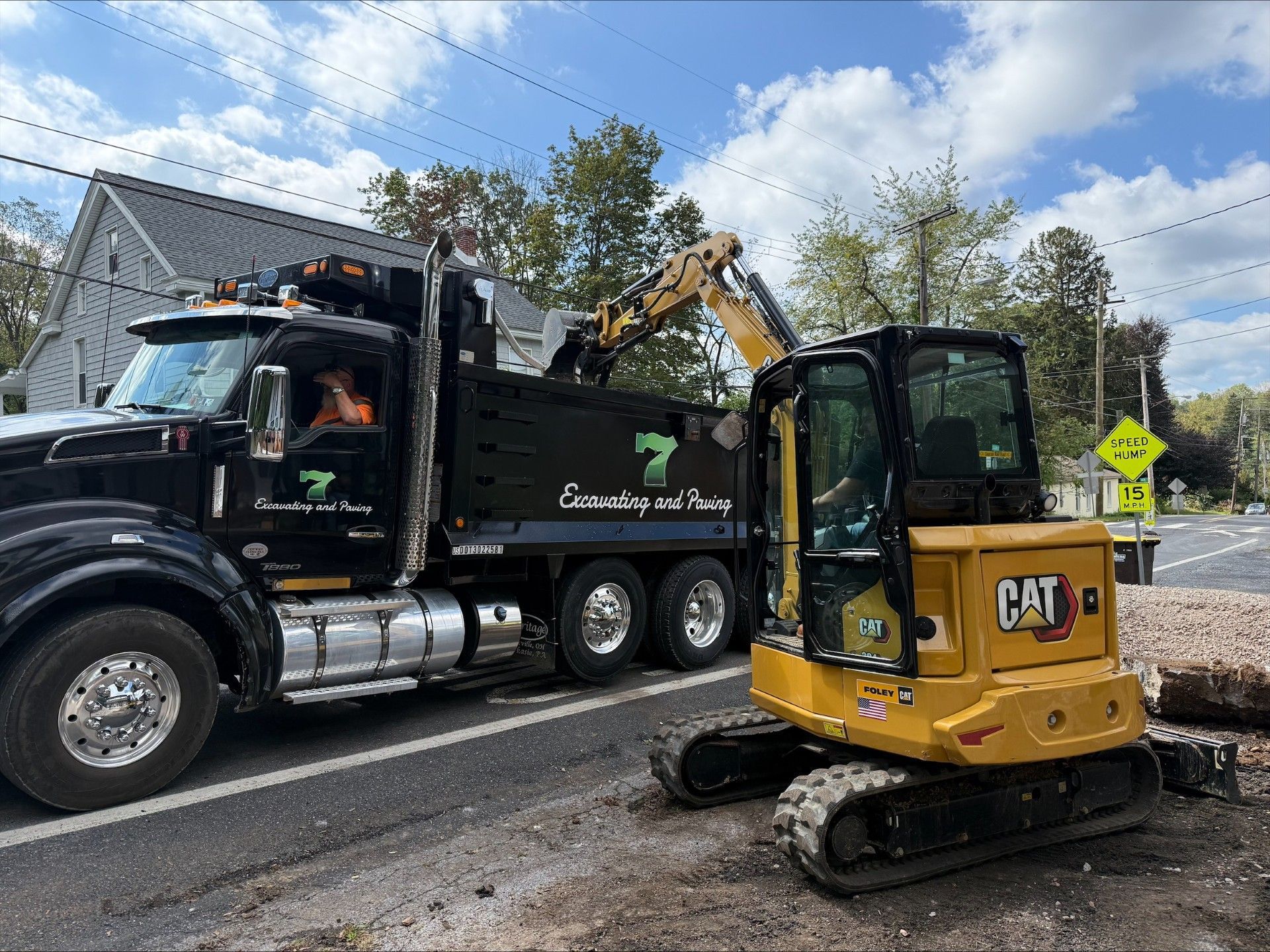 A small yellow CAT excavator loads a black dump truck on a roadside.