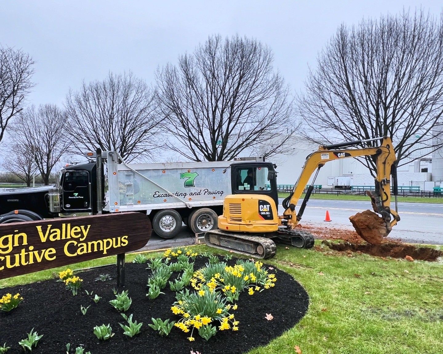Excavator and dump truck at a landscaped campus entrance with flowers and a sign on a rainy day