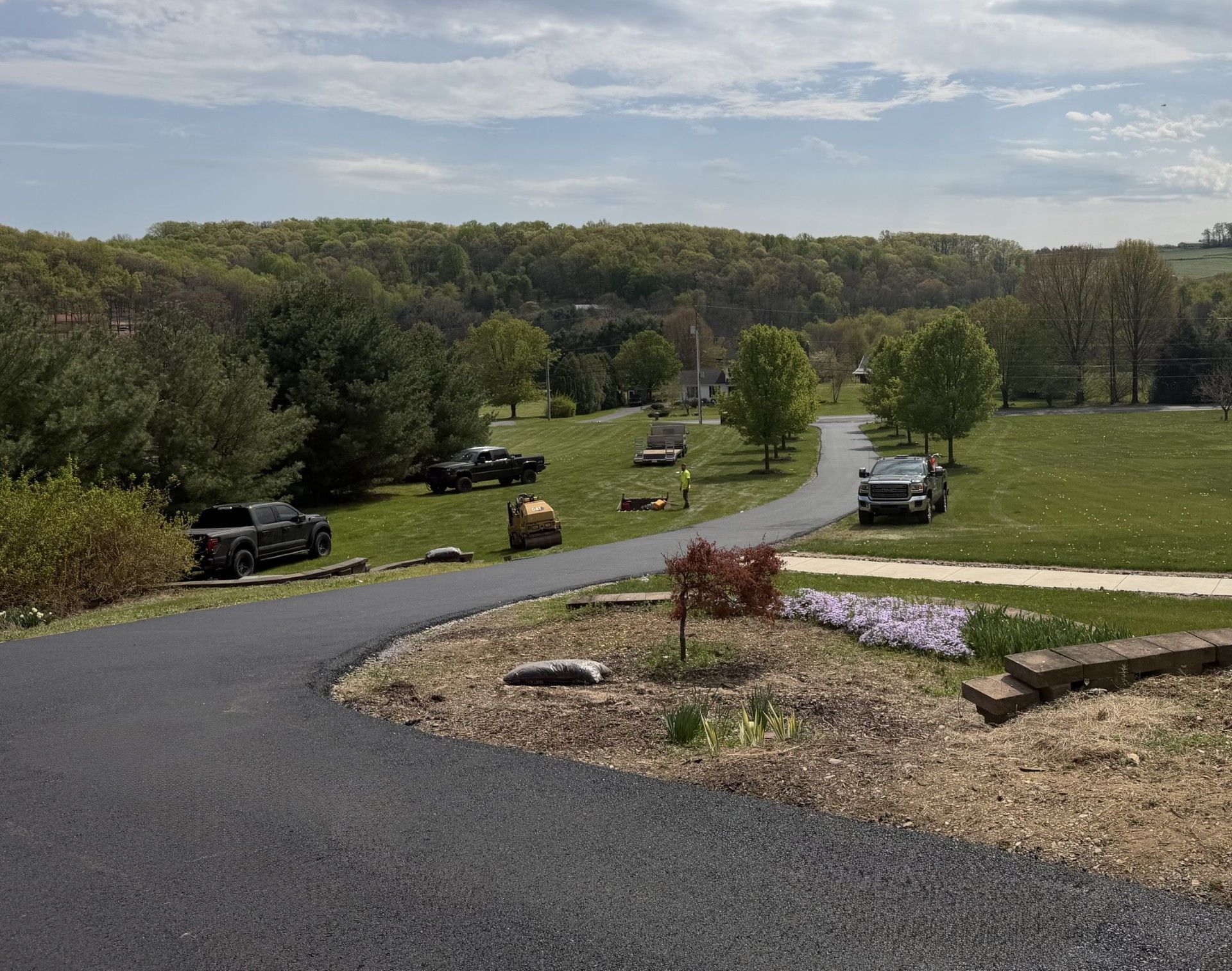 Curving driveway through a grassy rural landscape with parked vehicles and trees under a cloudy sky