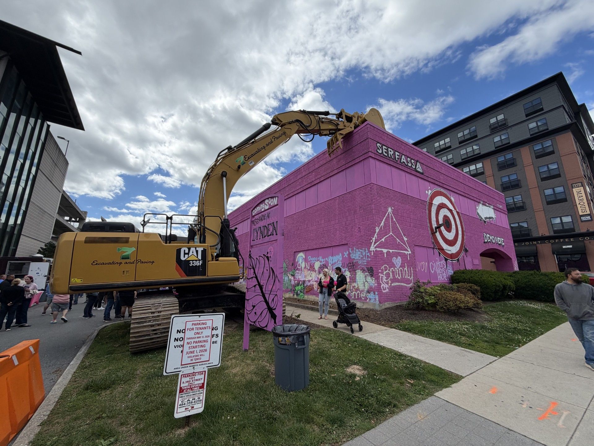 Yellow excavator lifting a giant pink cube structure on a city street with pedestrians nearby