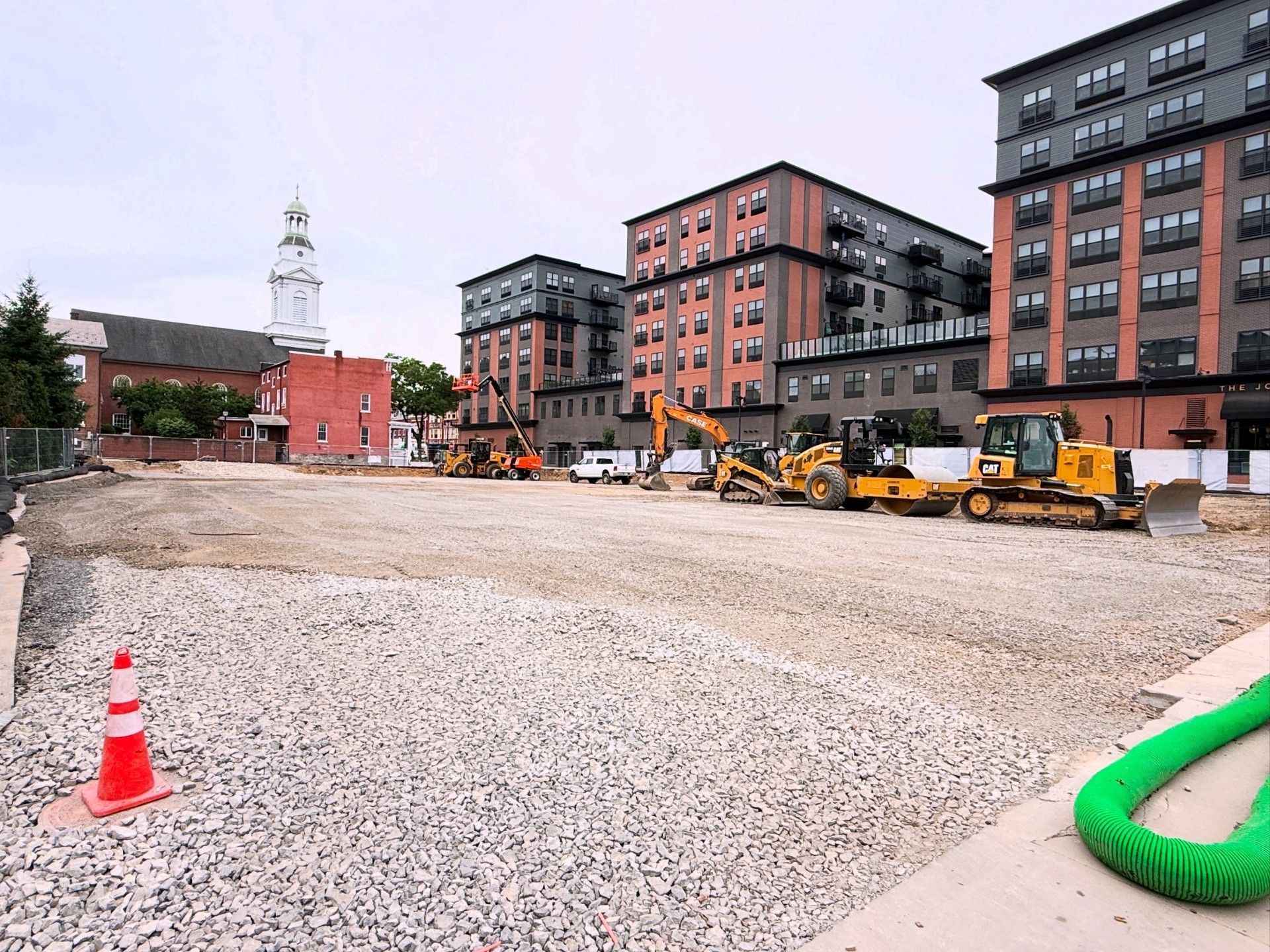 Construction site with gravel lot, orange excavator, and brick apartment buildings under overcast sky