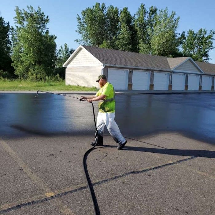 A worker in a high-visibility yellow shirt sprays dark asphalt sealer onto a parking lot surface.
