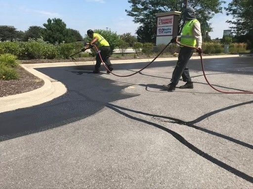 Two workers in neon safety vests use spray equipment to apply black sealant to an asphalt parking lot.