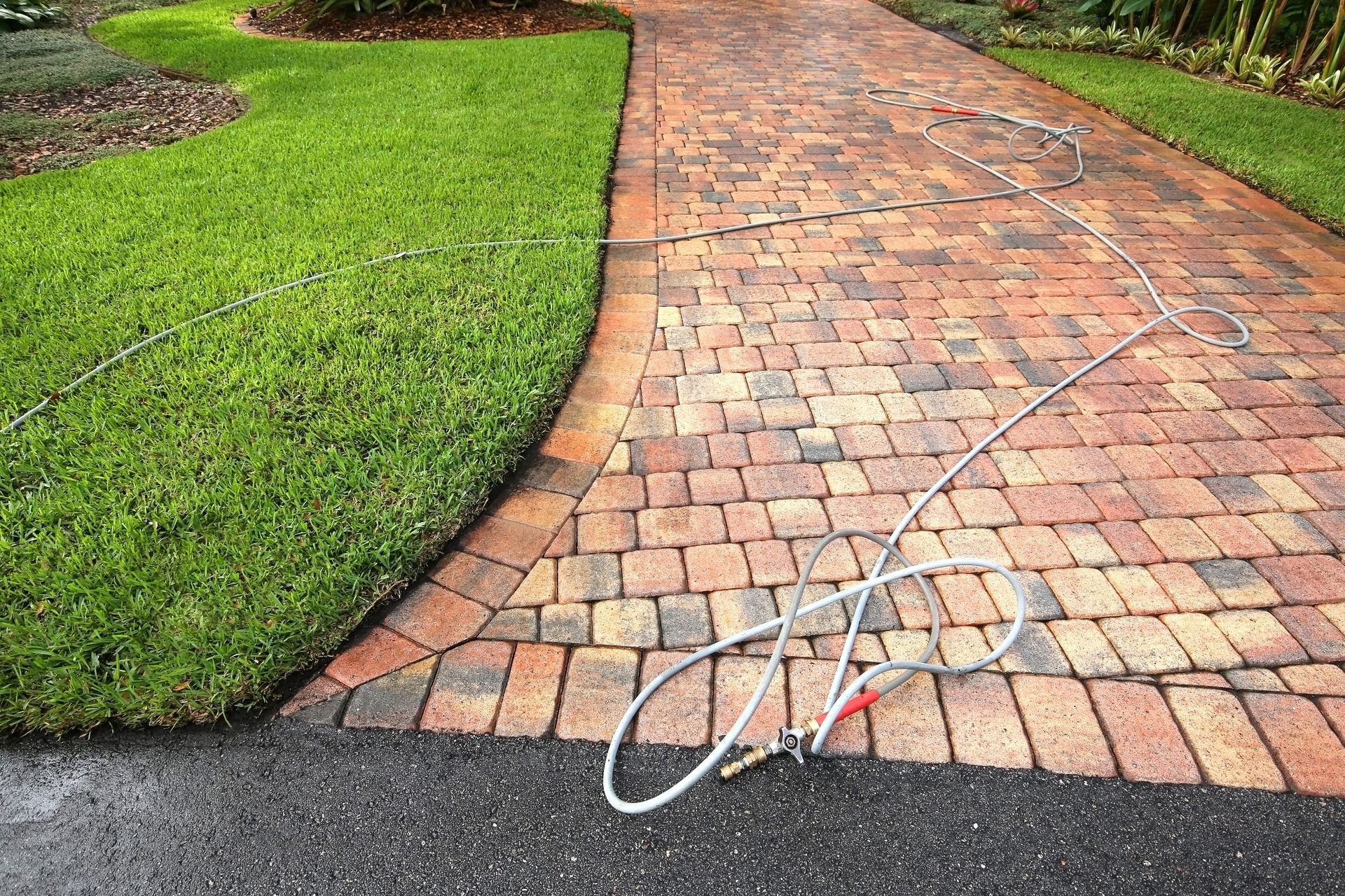 Brick paver walkway being cleaned with a hose, next to green lawn and asphalt driveway.