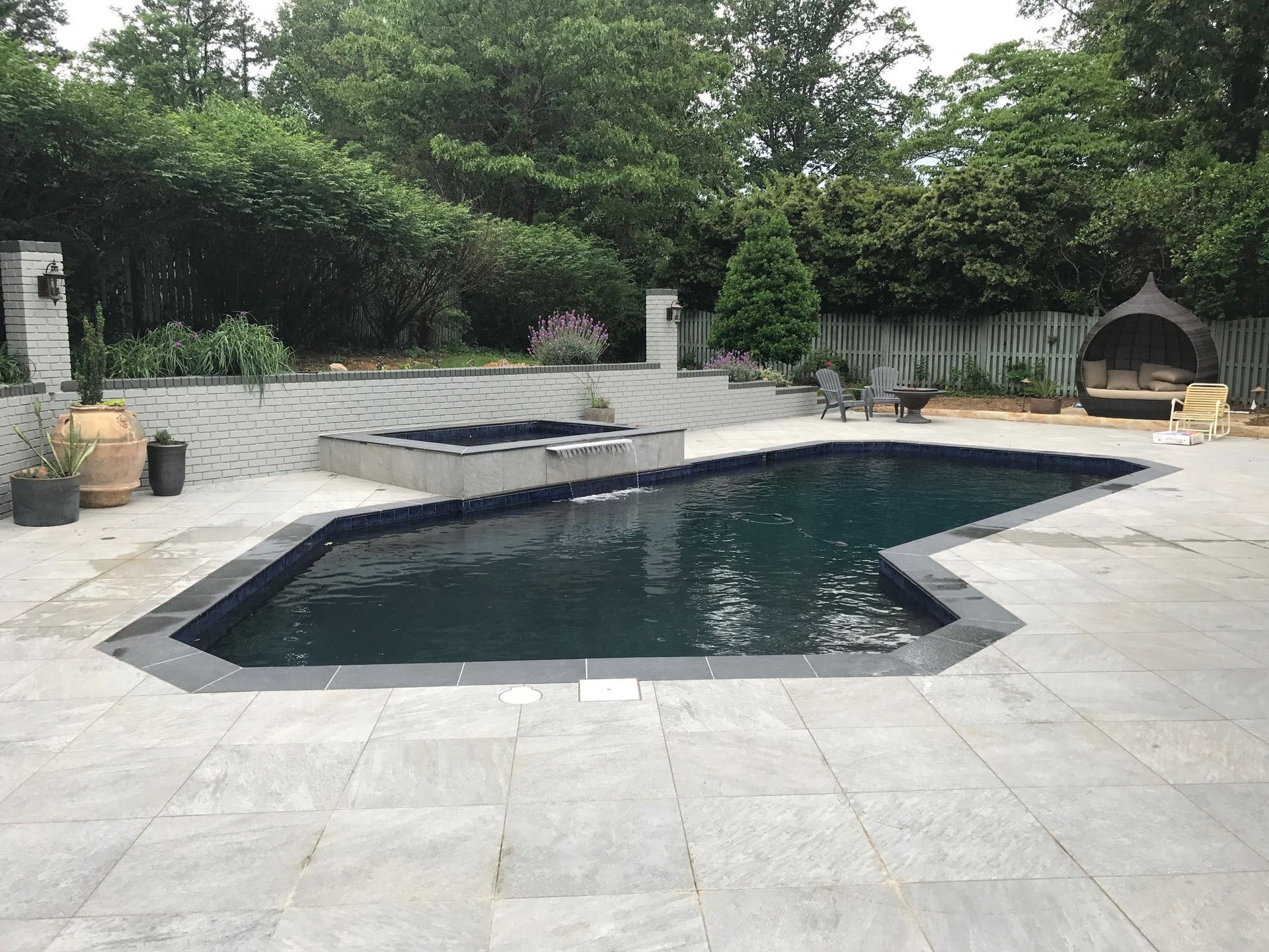 Backyard patio with an irregular swimming pool, gray stone deck, and outdoor fireplace surrounded by greenery