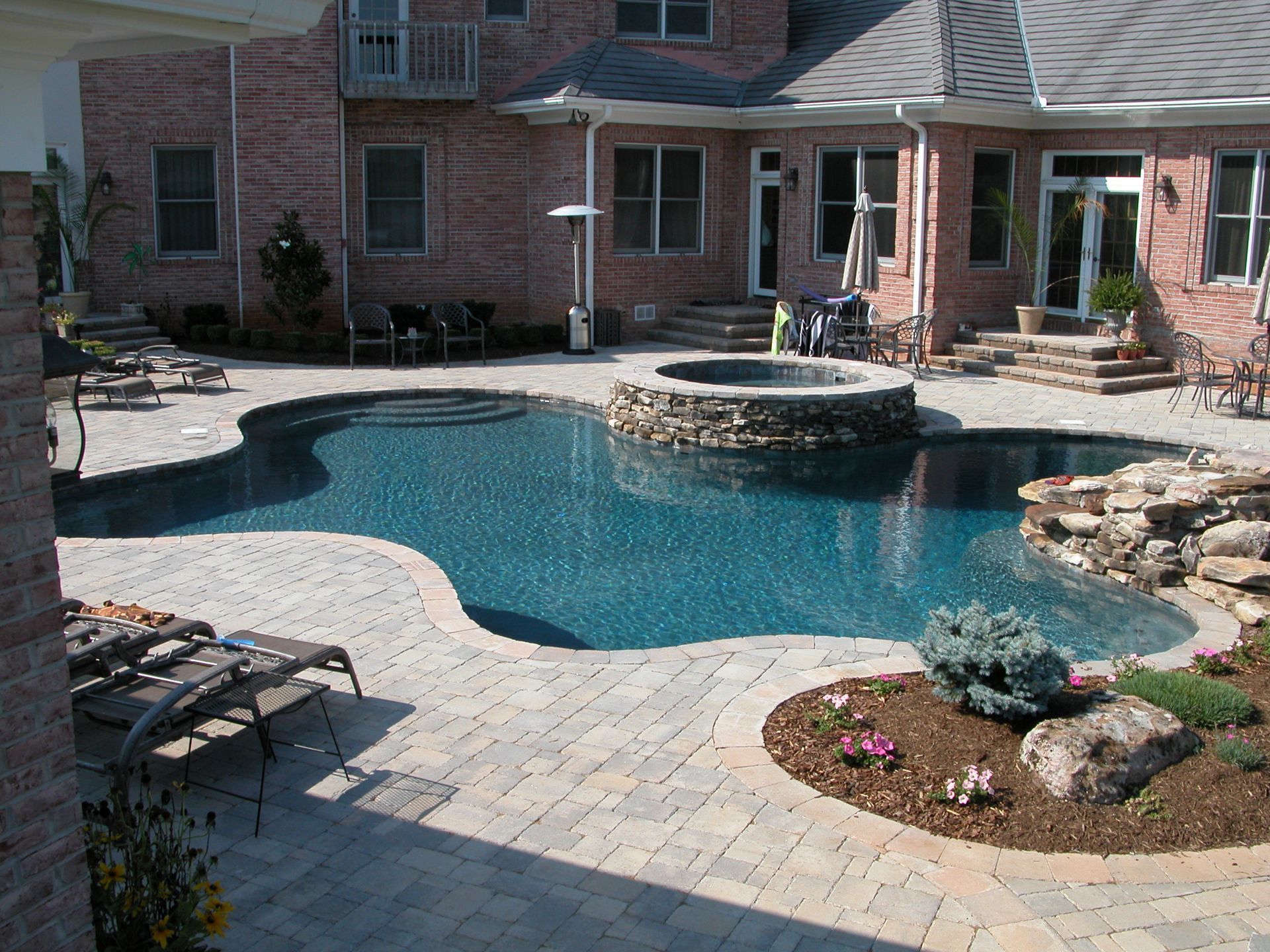 Backyard swimming pool with stone hot tub, brick patio, and lounge chairs beside a house