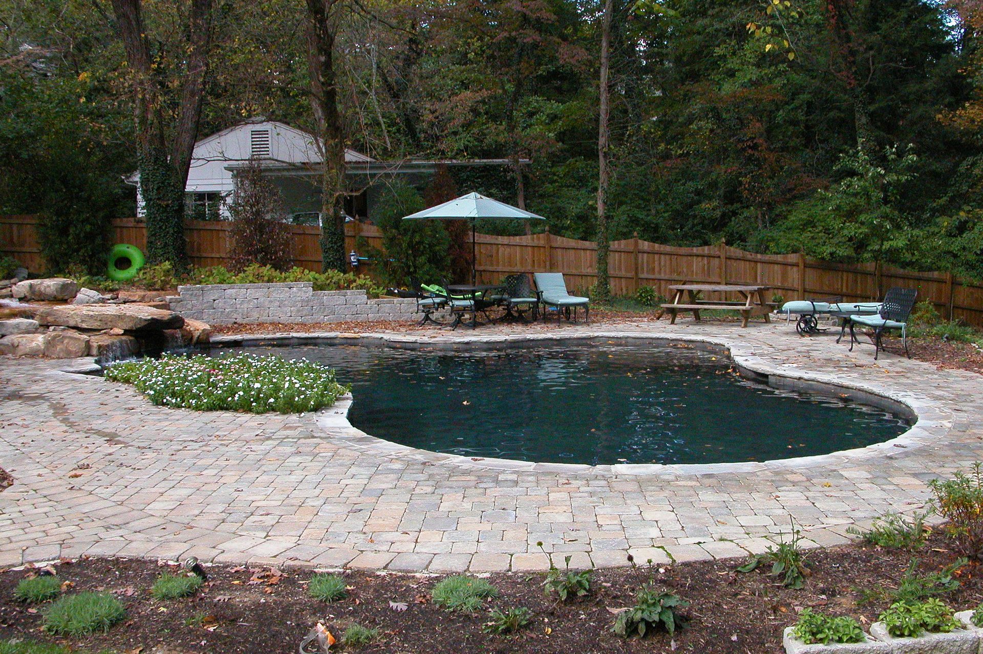 Backyard swimming pool with stone patio, lounge chairs, umbrella, and wooded garden surroundings
