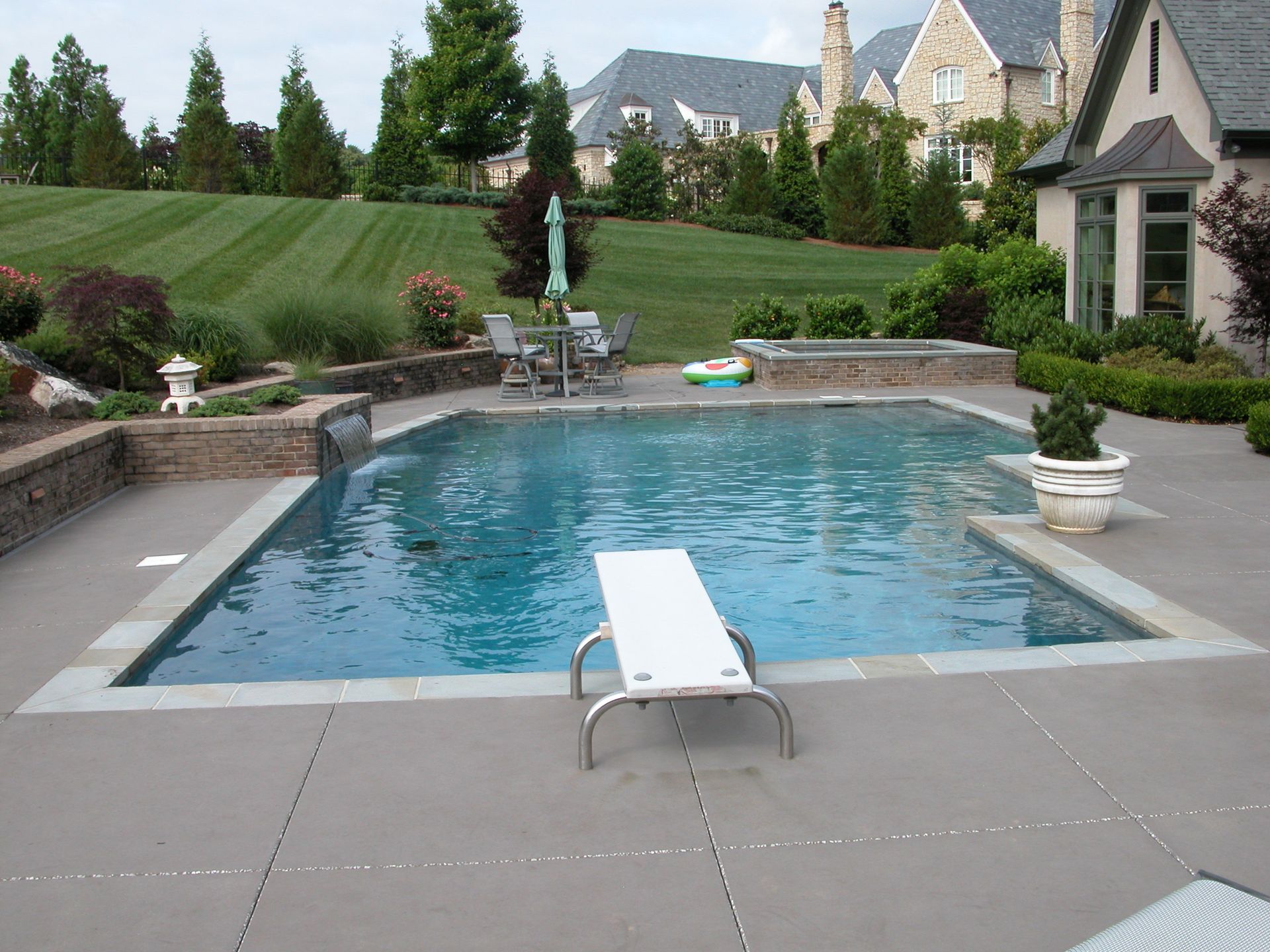 Backyard pool with lounge chair, stone patio, and landscaped lawn beside a house