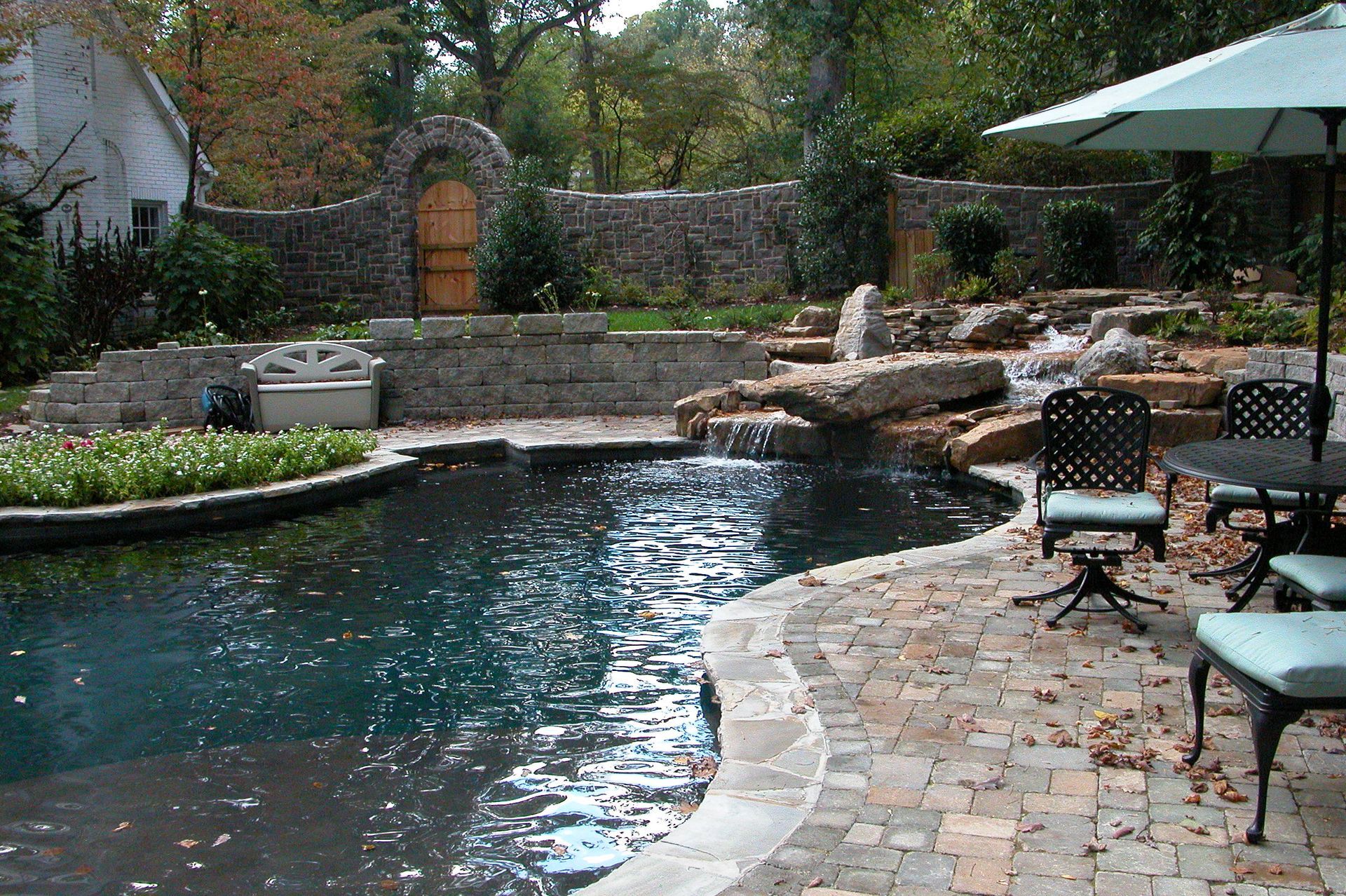 Backyard patio with curved swimming pool, stone deck, lounge chairs, and umbrella under trees