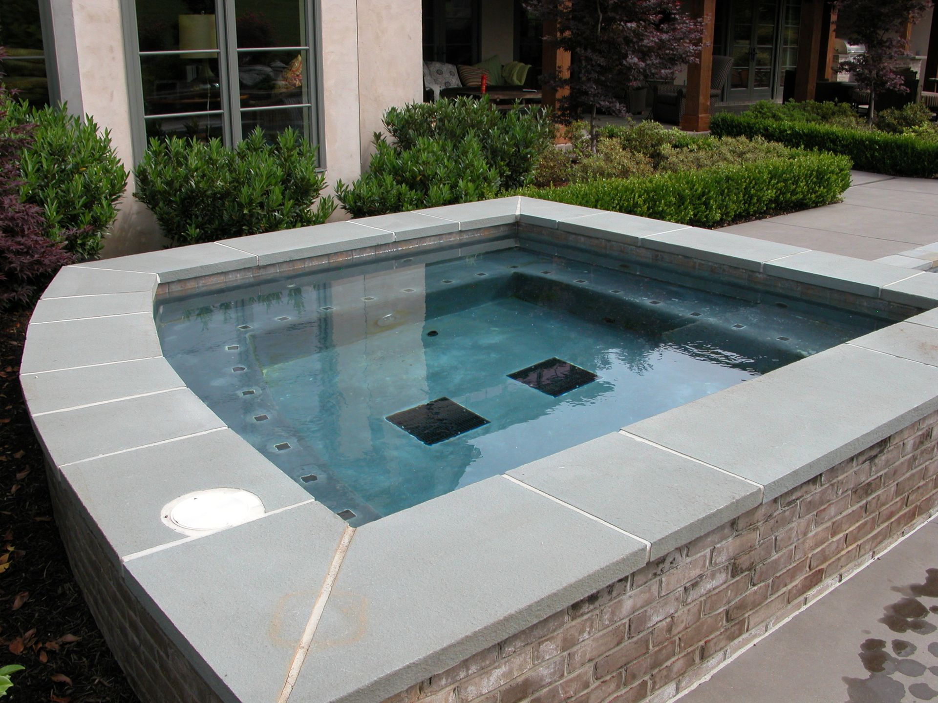 Small rectangular outdoor hot tub with clear water, gray stone coping, and brick sides beside a patio building