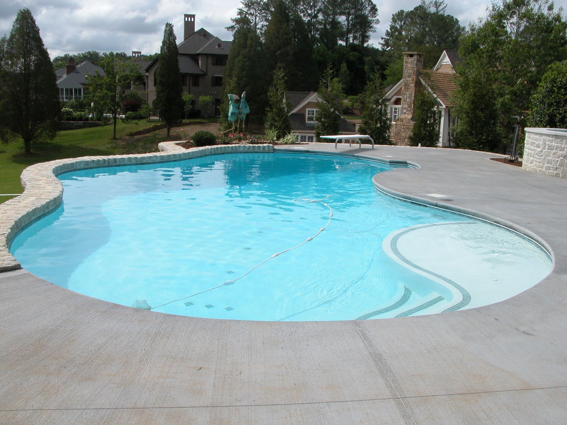 Backyard swimming pool with bright blue water and a stone deck, surrounded by trees and houses
