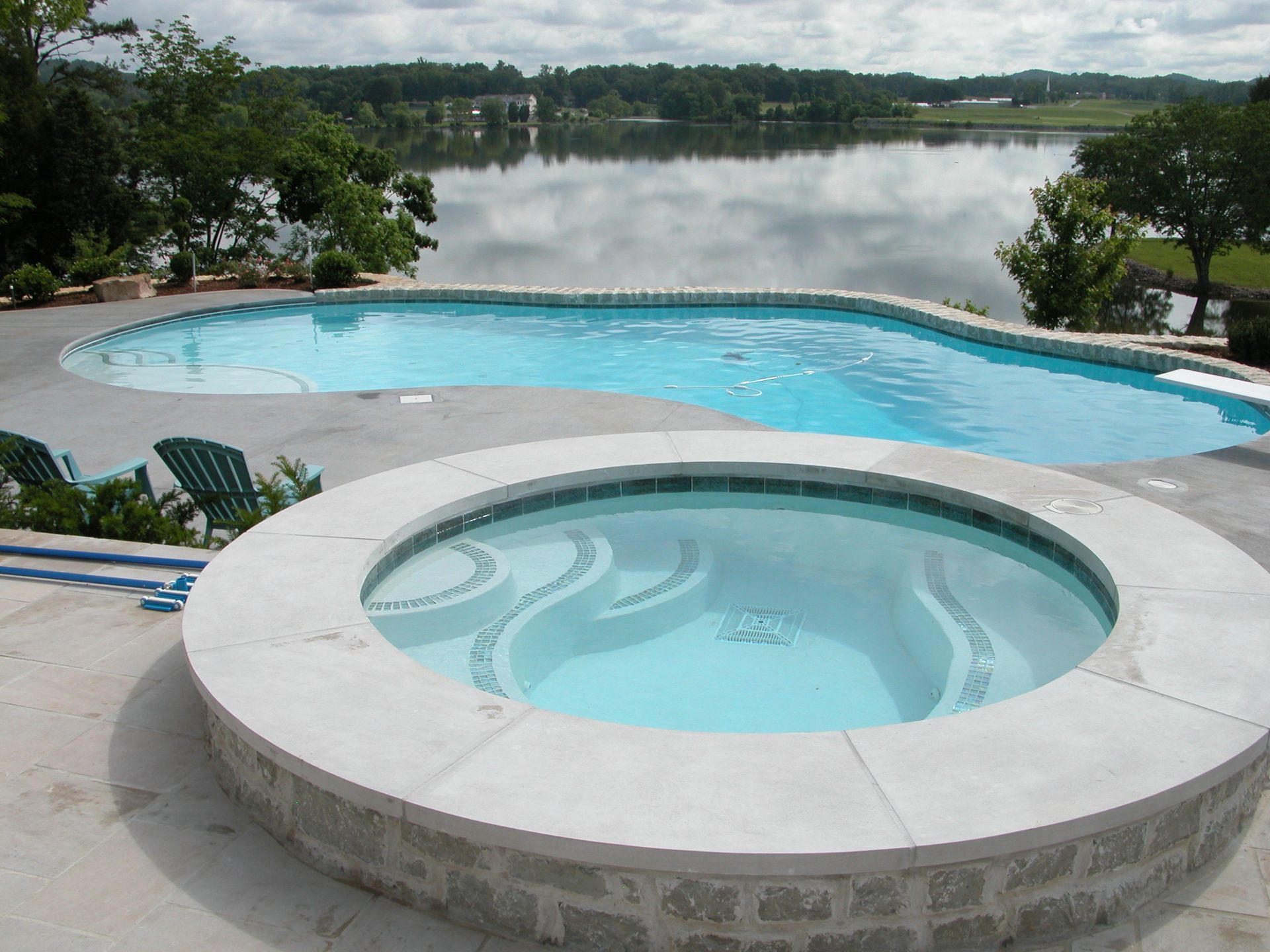 Patio with round hot tub and curved pool overlooking a calm lake and trees