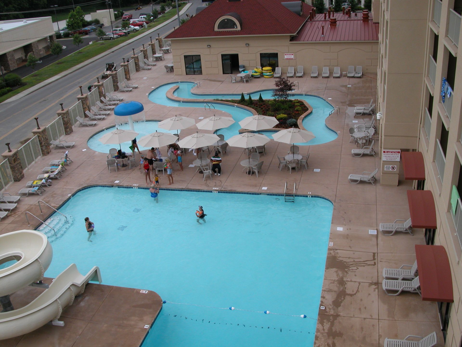 Hotel pool deck with blue pools, white umbrellas, lounge chairs, and a few people swimming