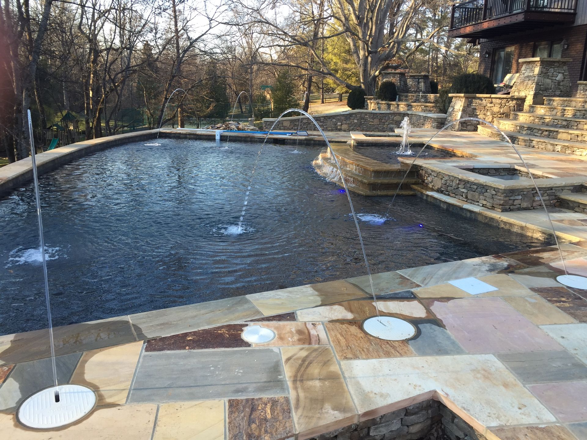 Backyard pool with fountains, stone patio, and house deck in the background