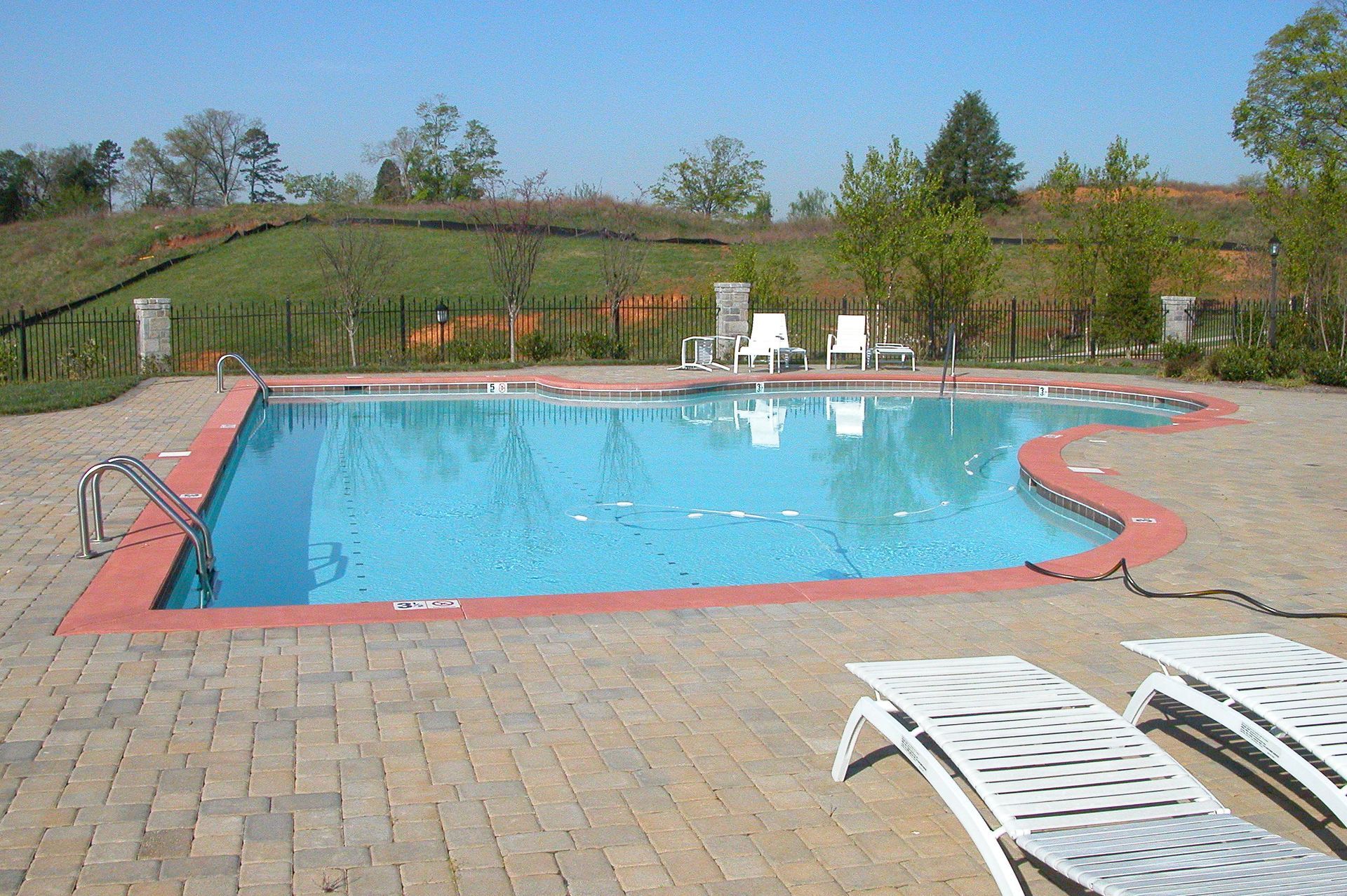 Outdoor swimming pool with blue water, white lounge chairs, and a brick patio in a grassy landscape